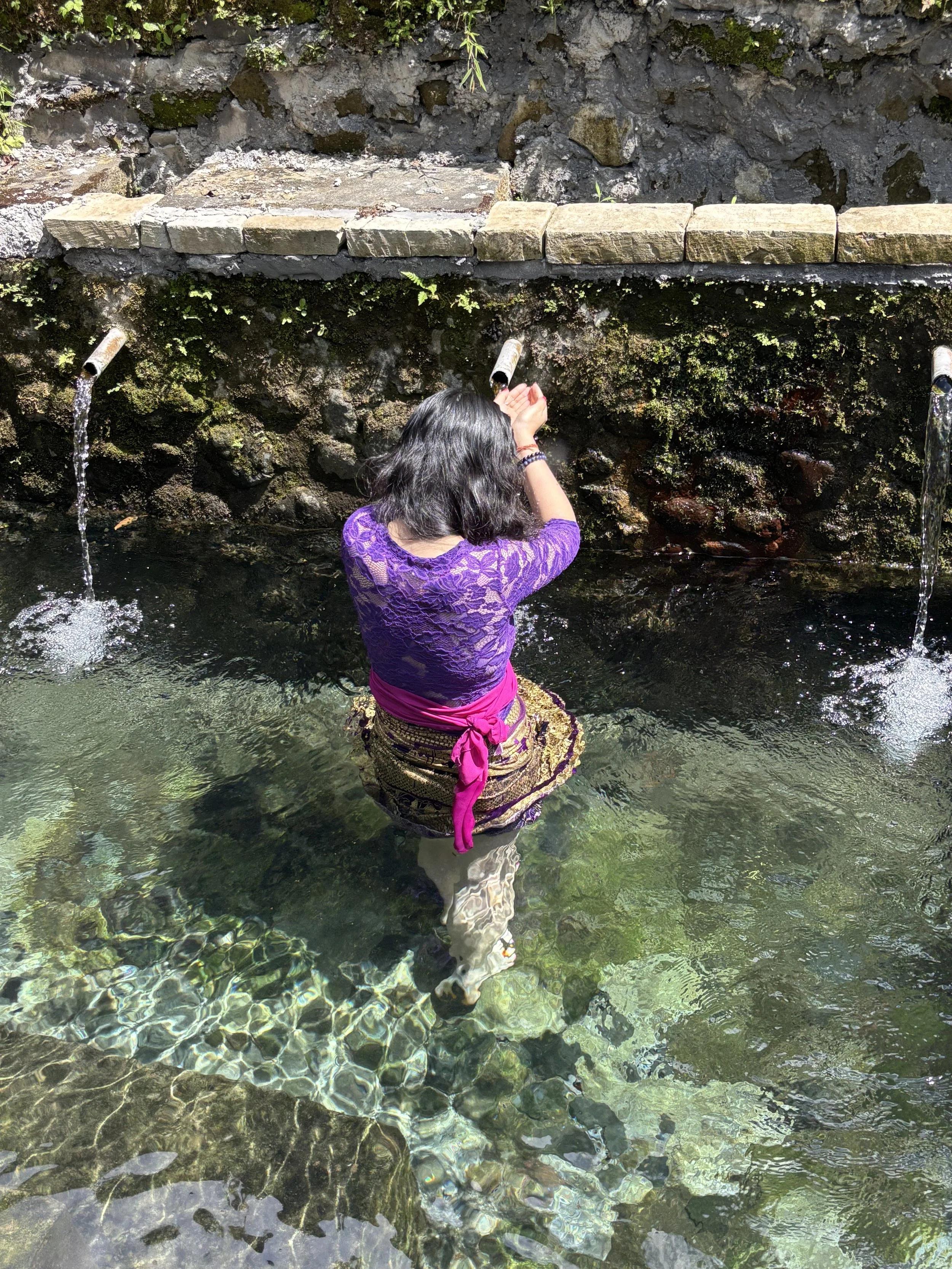 A woman standing in a shallow, clear water stream with a rocky wall in the background. She is wearing a purple top and patterned skirt, facing away from the camera, with her hands pressed against an oral pipe for the water.