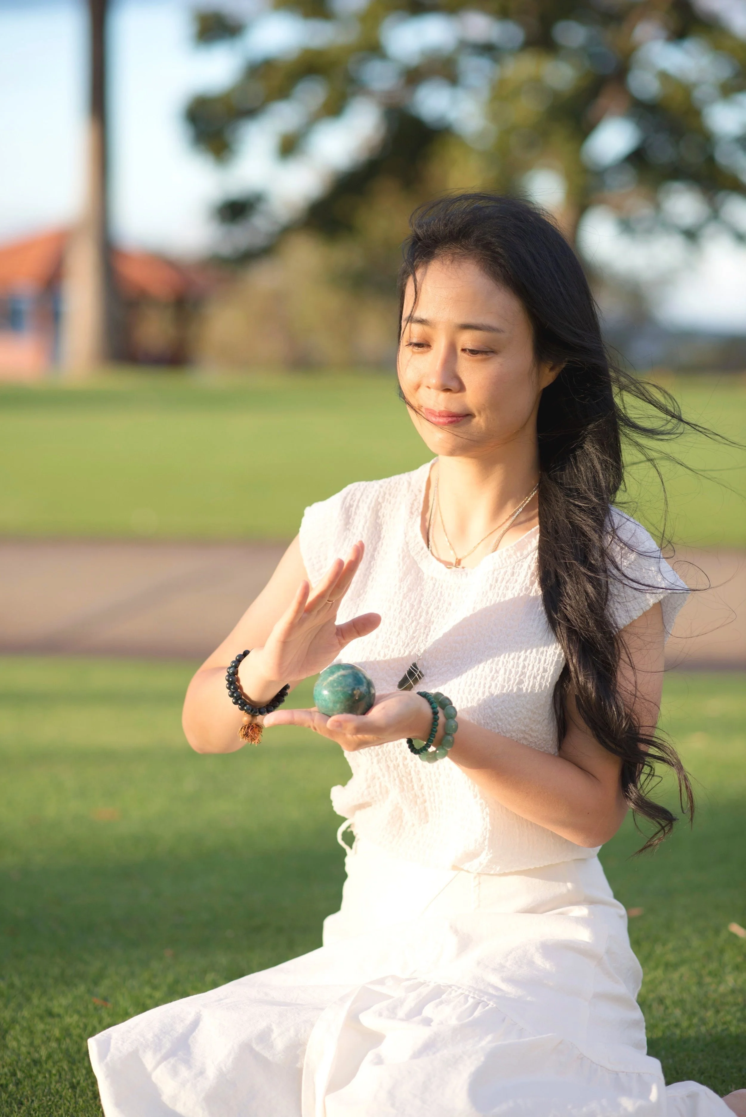 A woman meditating outdoors on grass, holding a stone sphere, with her eyes closed and peaceful expression.