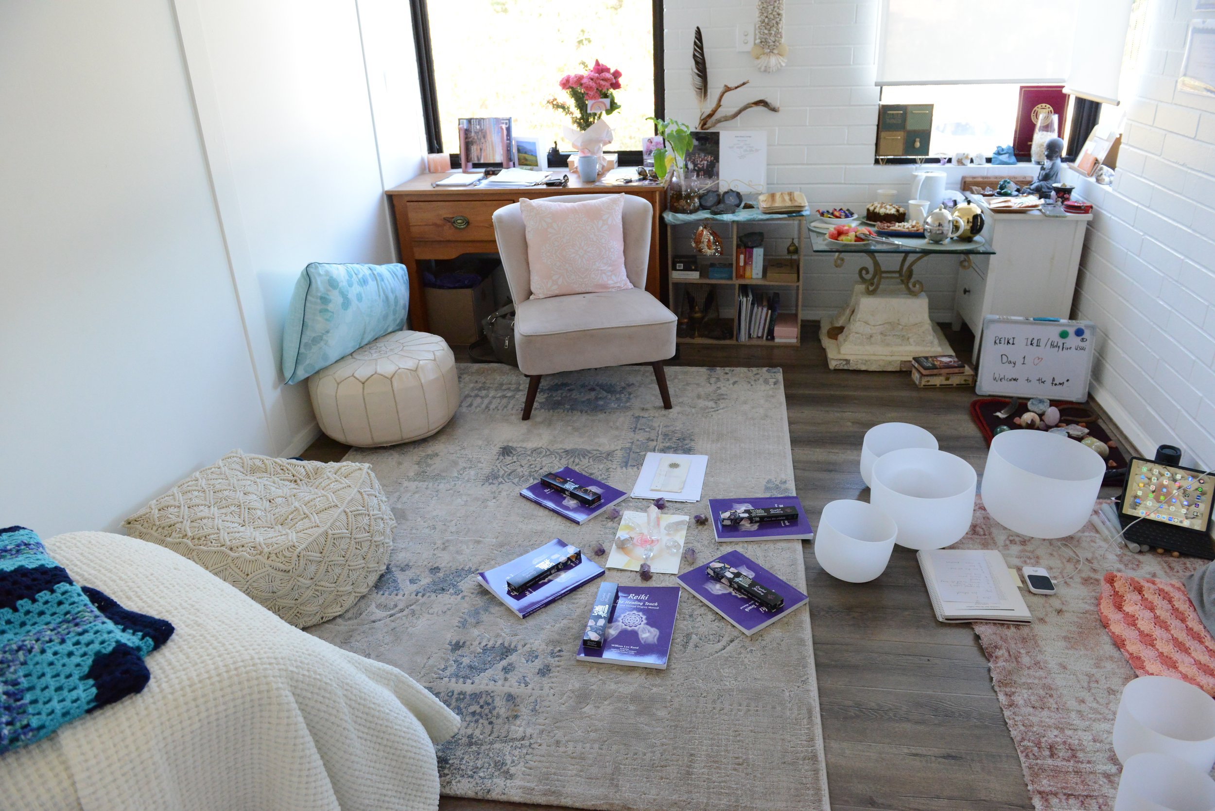 A cozy room with white walls and a window letting in natural light. The room has a white quilt with colorful crocheted blankets, a cream-colored armchair with a pink pillow, and a round ottoman. Stuffed animals and books are scattered on the floor an