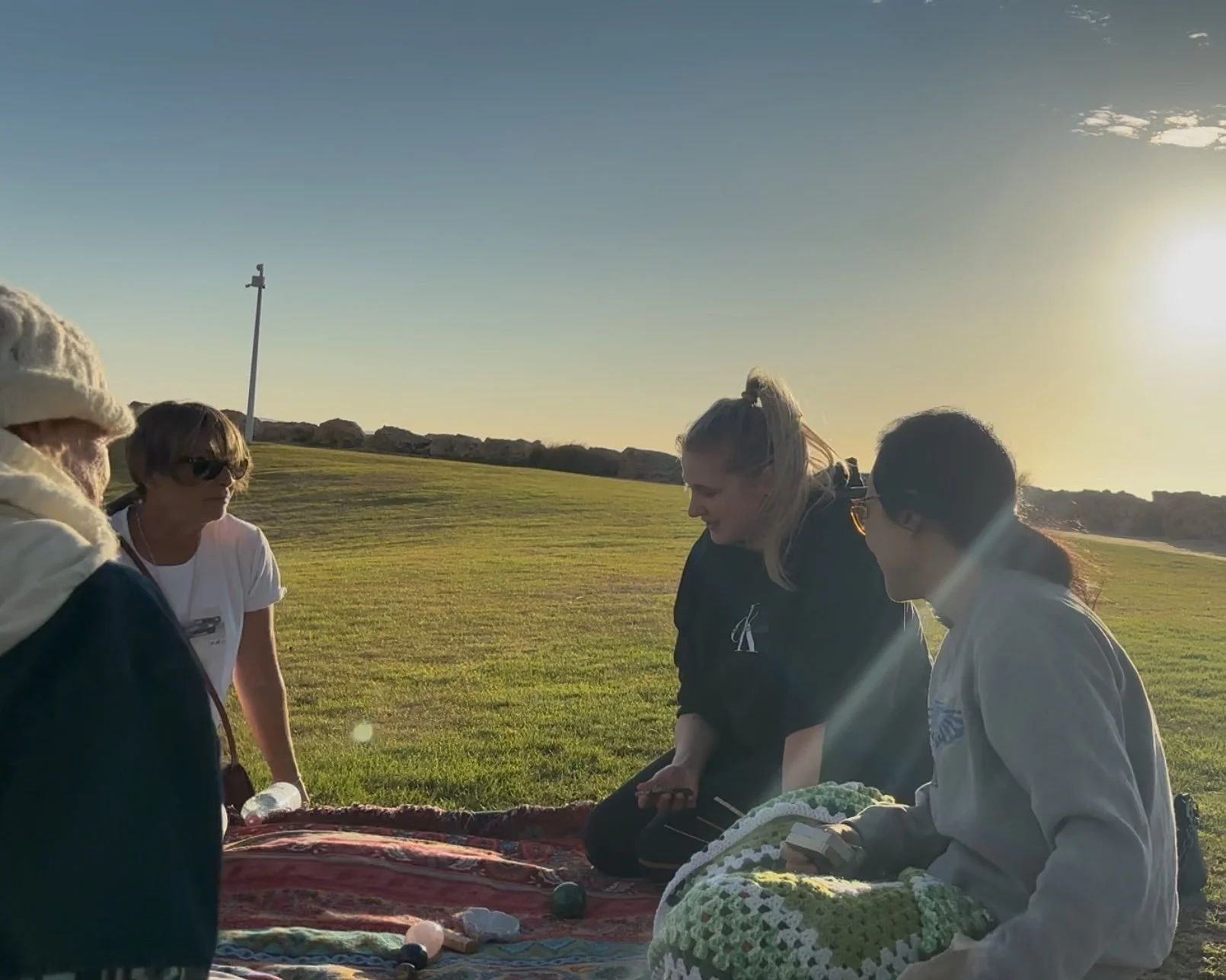 A group of women sitting and talking outdoors on a grassy field during sunset, with rocks and a light pole in the background.
