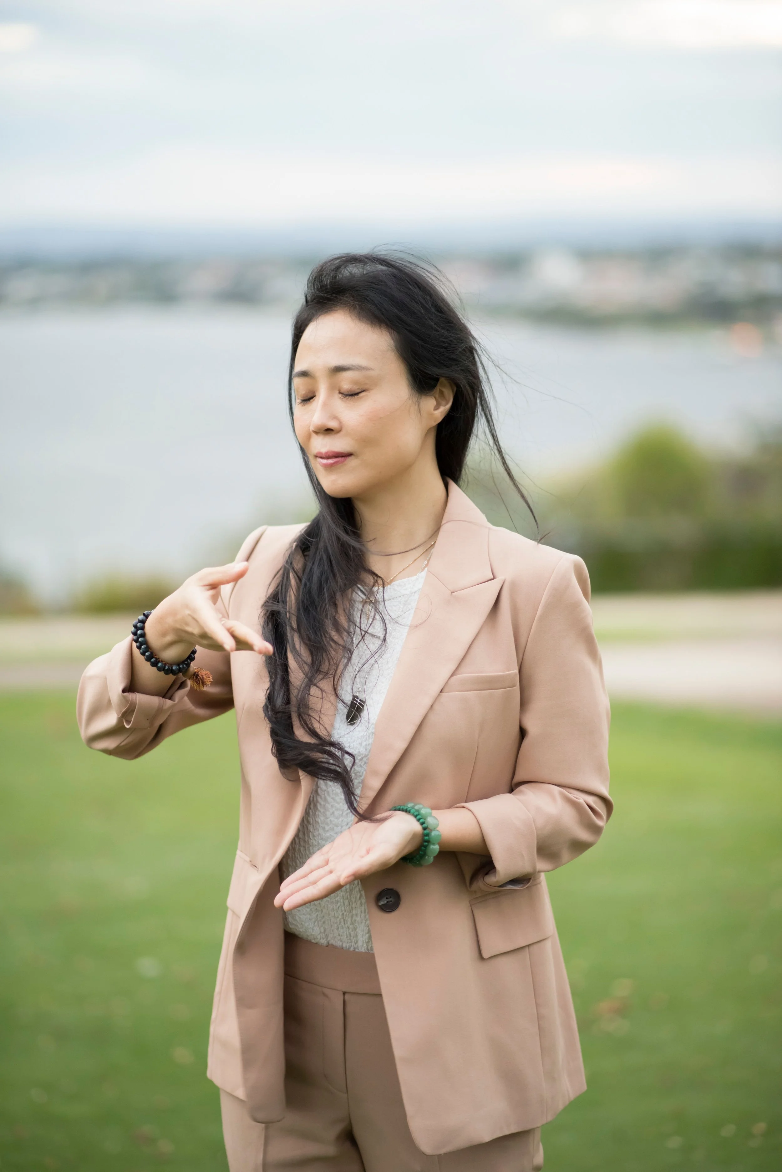 A woman with long black hair, wearing a beige suit and multiple bracelets, standing outdoors with eyes closed, against a blurred background of water and greenery.