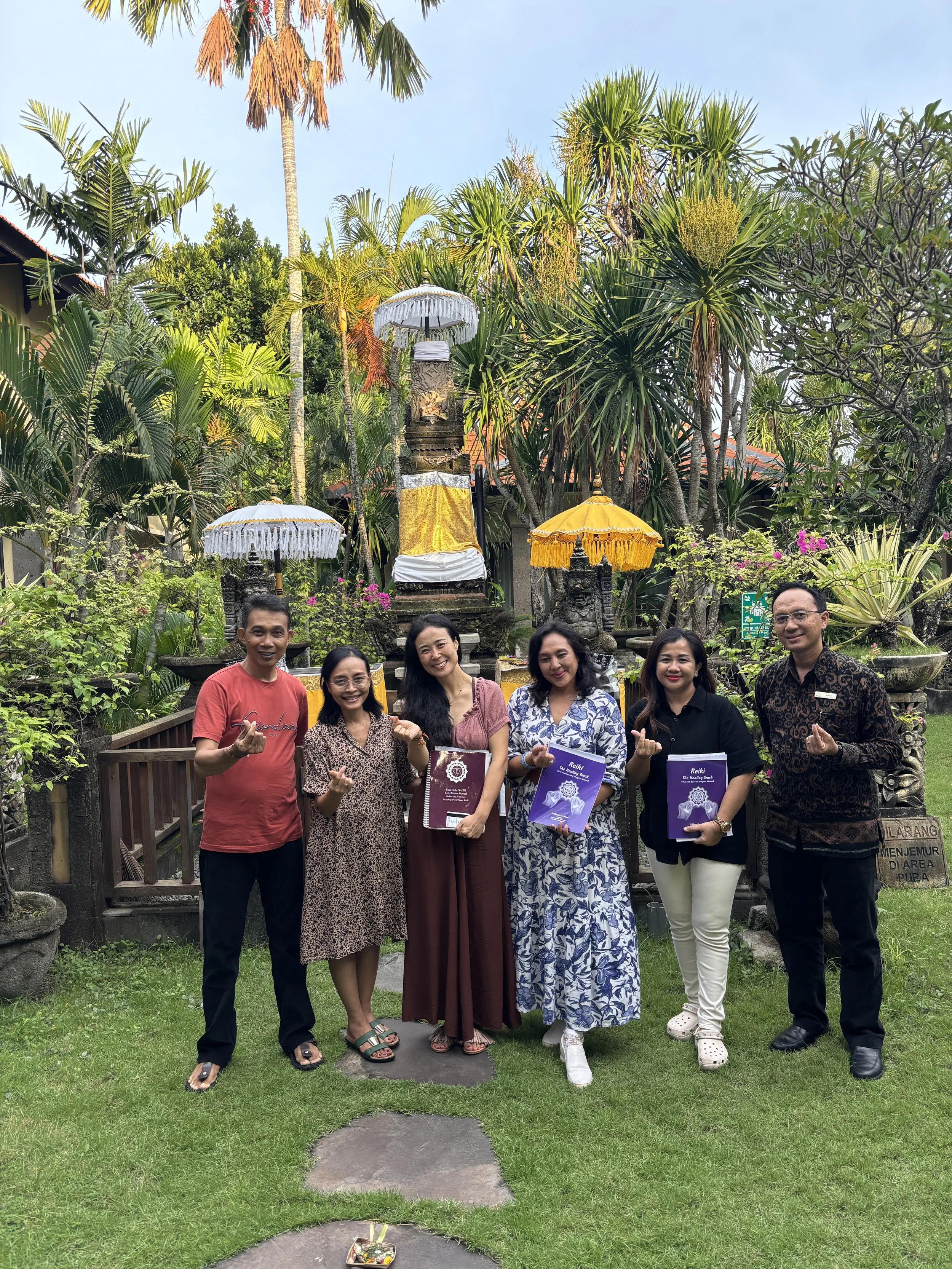 A group of seven people, five women and two men, standing in a lush garden with tropical plants and Balinese umbrellas, holding books and making a thumbs-up gesture.