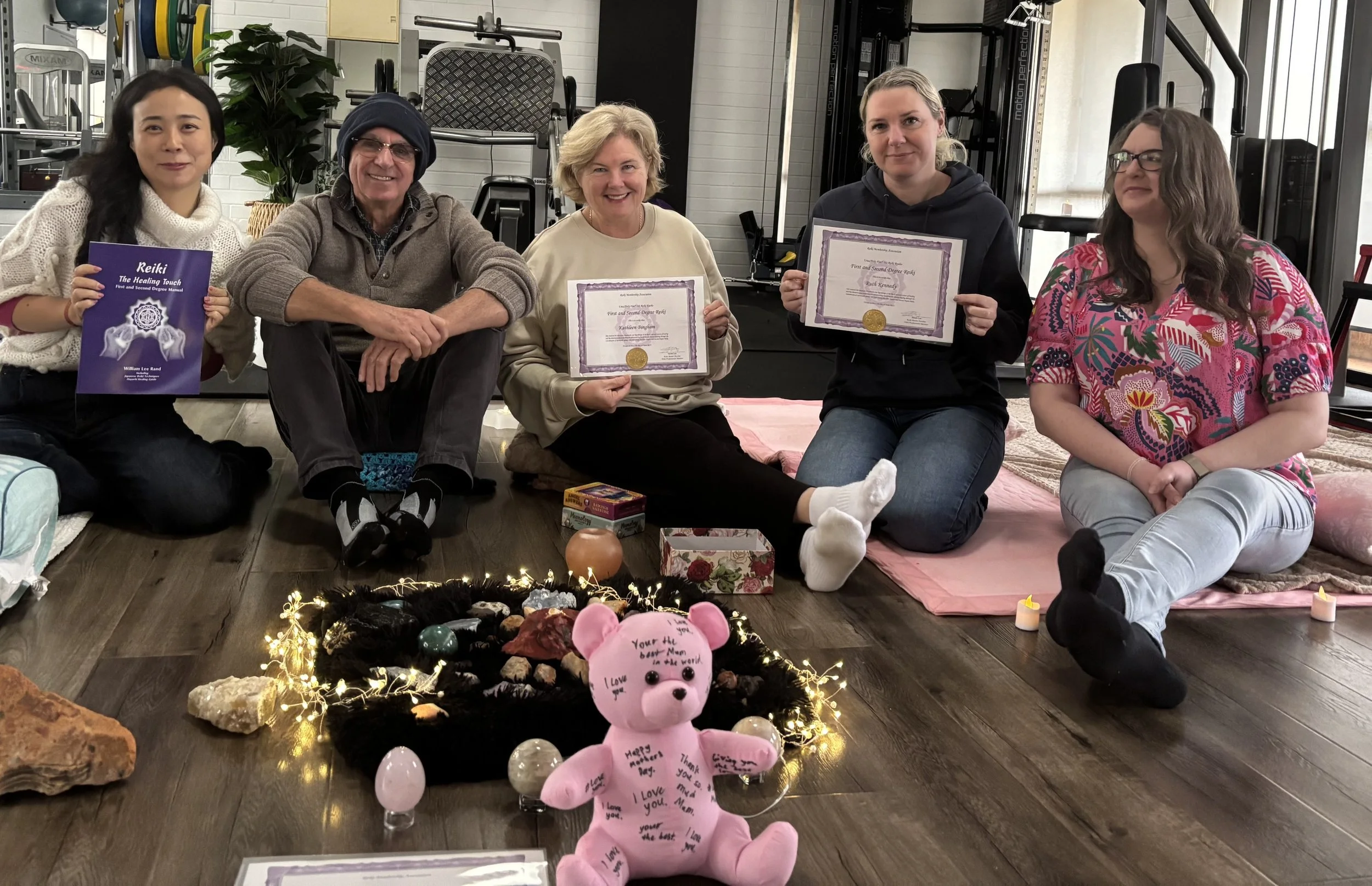 Group of five women sitting on the floor at a wellness or yoga space, holding certificates and a book titled "Reiki: The Healing Touch." In front of them is a small decorated area with a pink stuffed pig, candles, rocks, and fairy lights, celebrating