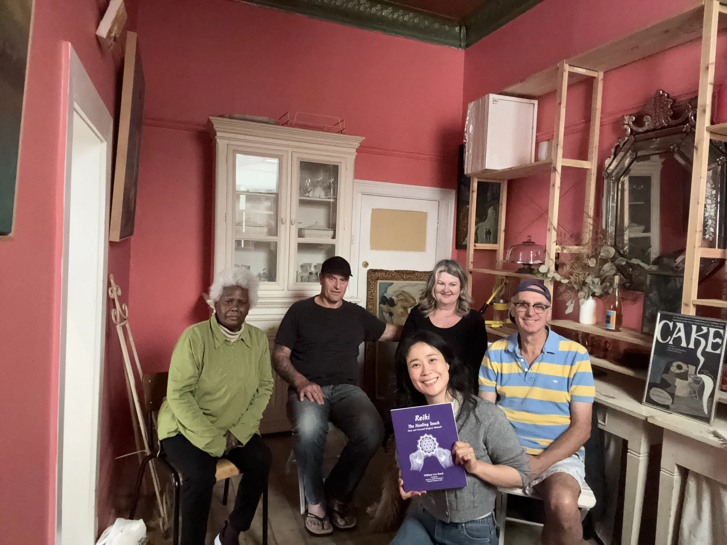 A group of seven people, four men and three women, posing inside a room with pink walls and wooden shelving. One woman in the front is holding a purple book titled 'Reiki The Healing Touch'. They are smiling and standing close together, appearing to 