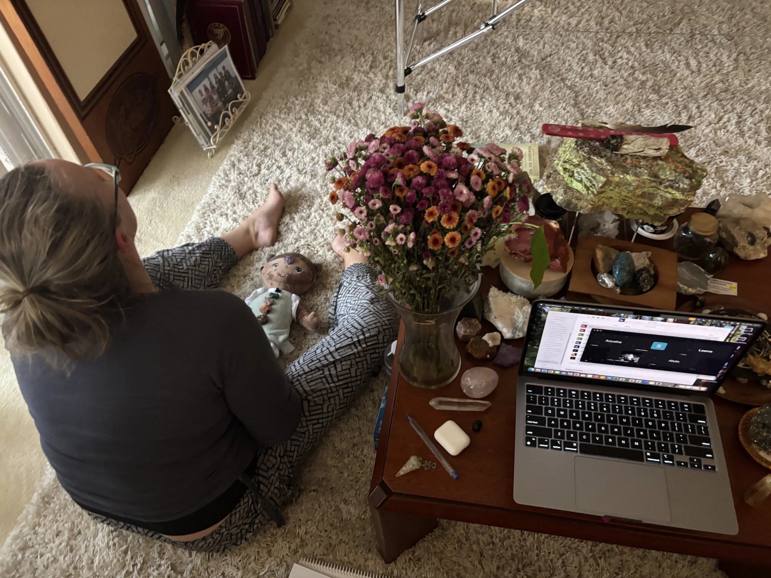 A person with long blonde hair and glasses, dressed in a dark blazer and patterned pants, kneeling on a beige carpet, with a flower arrangement, a doll, and various objects on a wooden table nearby. There is a laptop open with multiple application wi