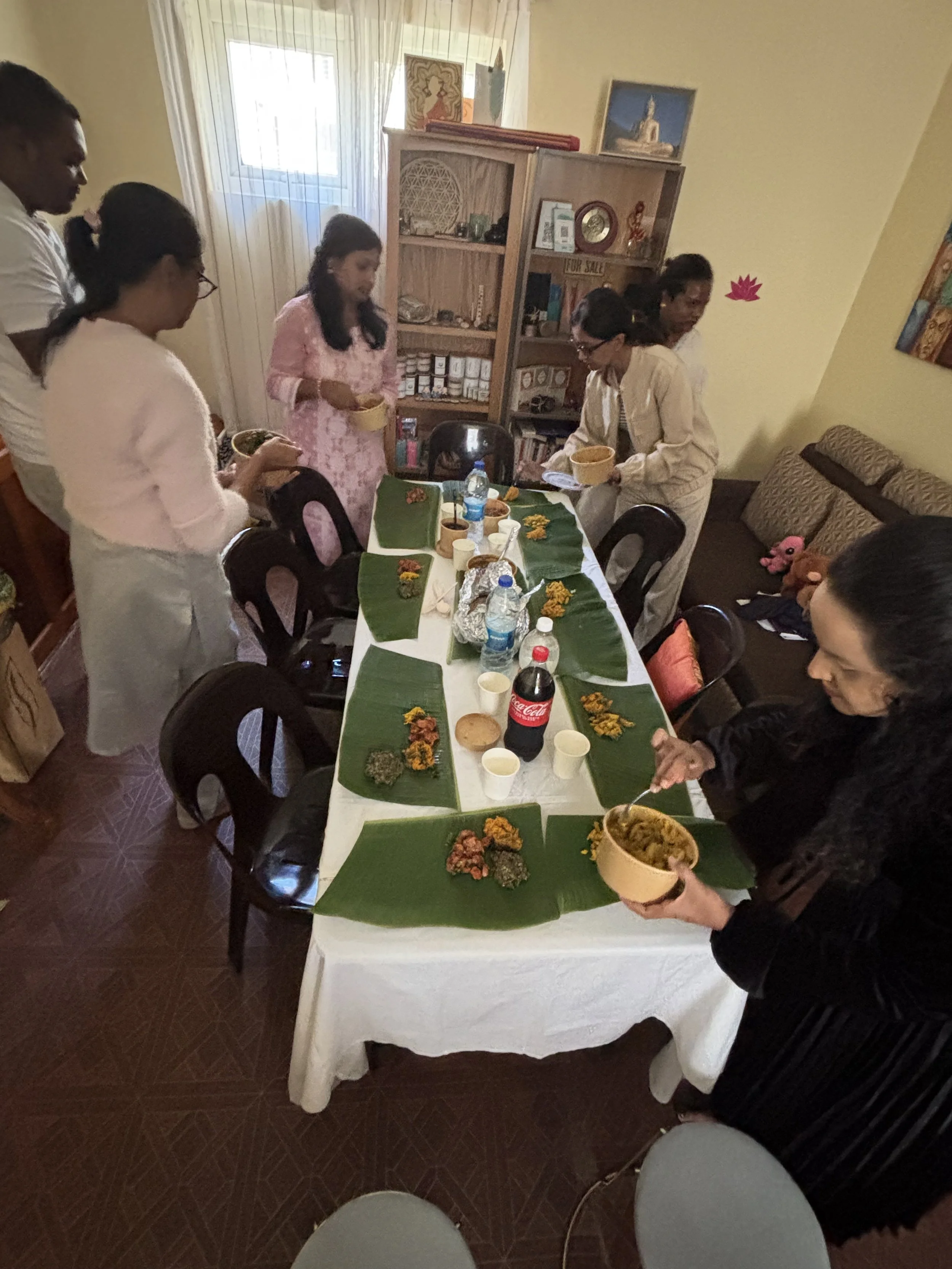 Group of people gathered around a table with Indian food and drinks, some serving and others eating, in a cozy indoor setting.