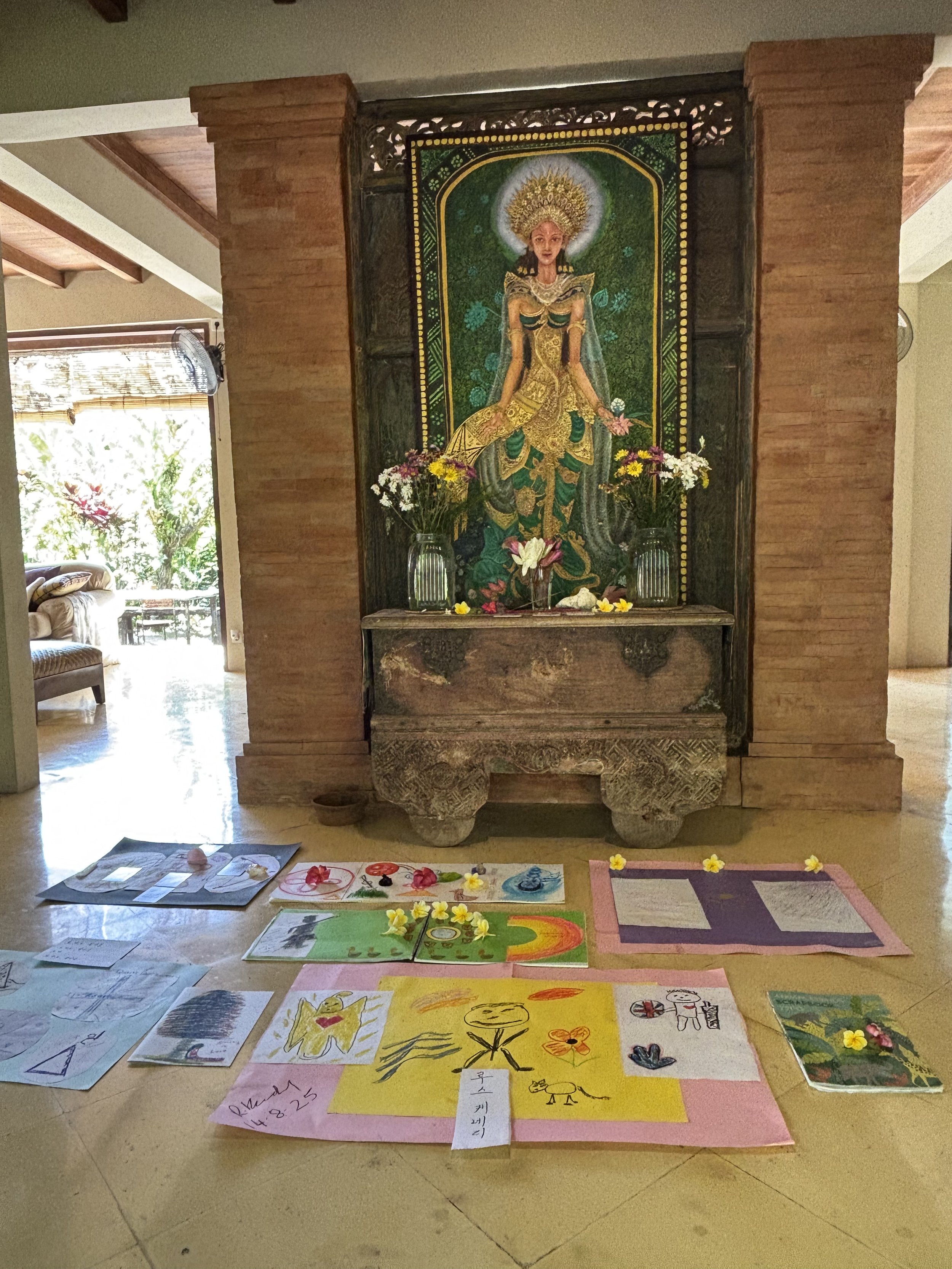 A religious altar with a colorful framed religious painting and flowers, in a room with windows and furniture, and children’s drawings and artwork laid out on the floor in front.