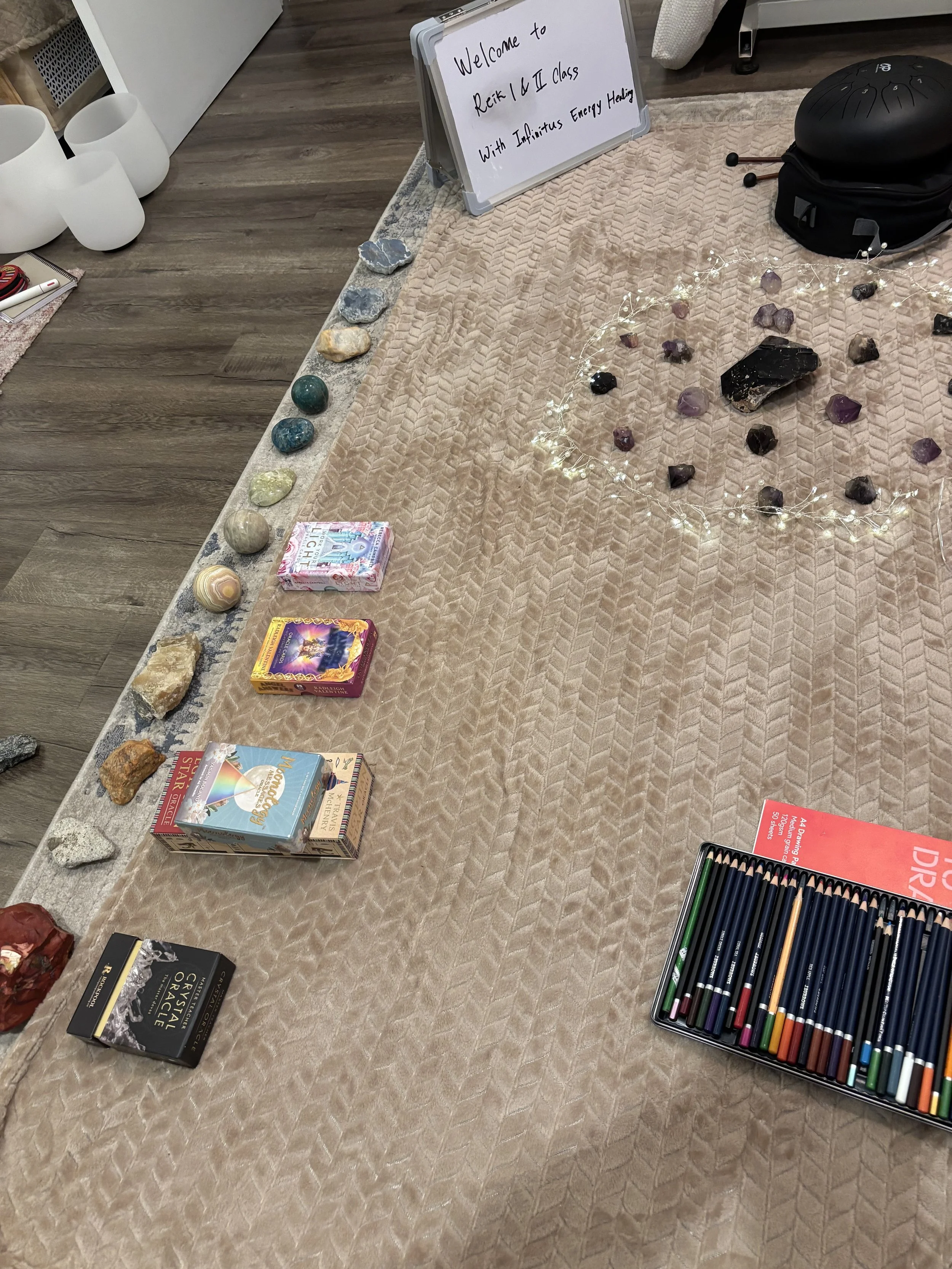 Various items displayed on a beige textured cloth, including a row of gemstones, boxes of art supplies, some books, a black musical instrument case, and a whiteboard with a welcome message for a Reiki and energy healing class.