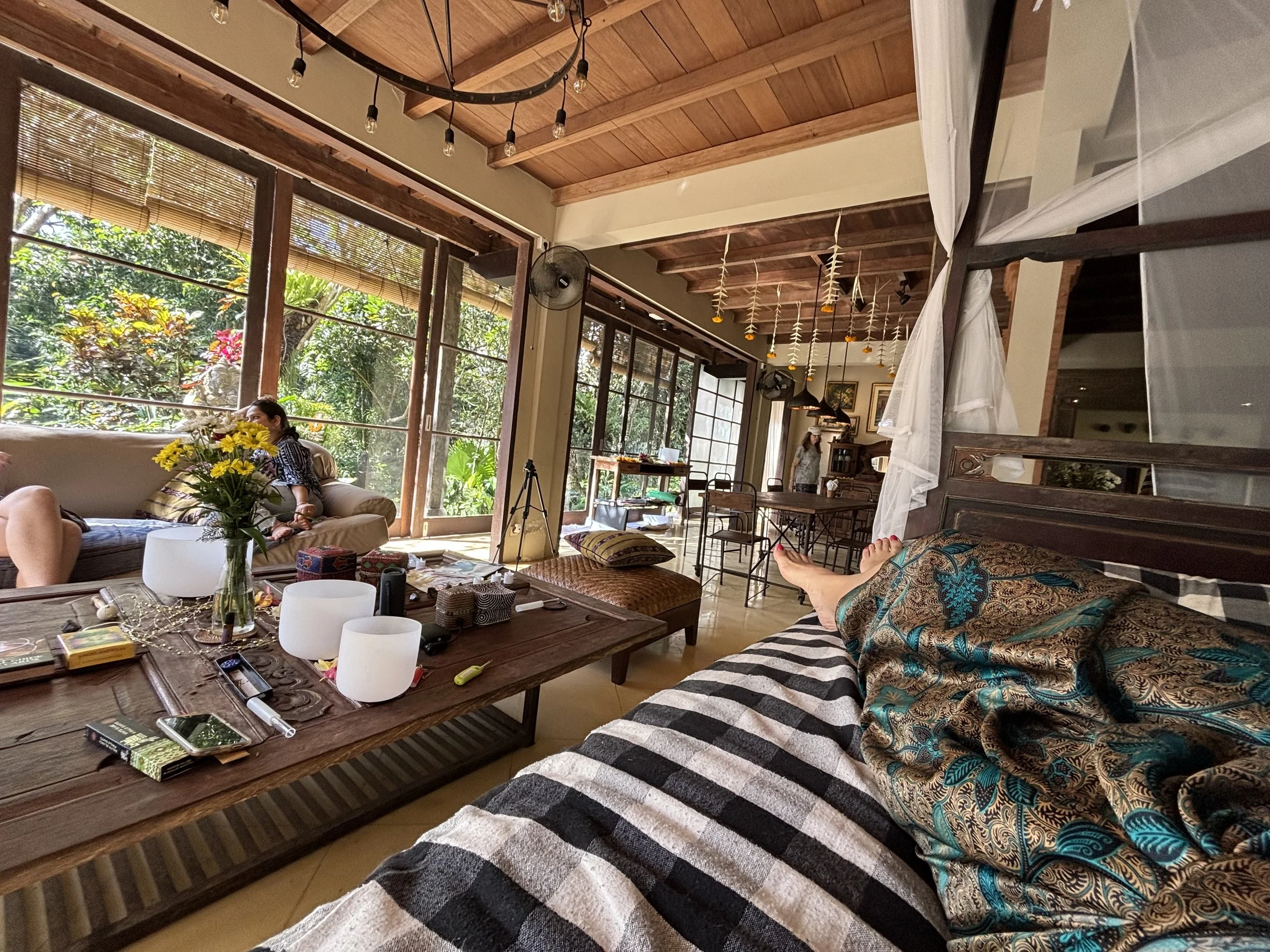 Interior of a bright, airy living space with large windows, wooden ceiling, and casual seating. There are plants outside, and various items including flowers, books, and electronic devices on a wooden table.