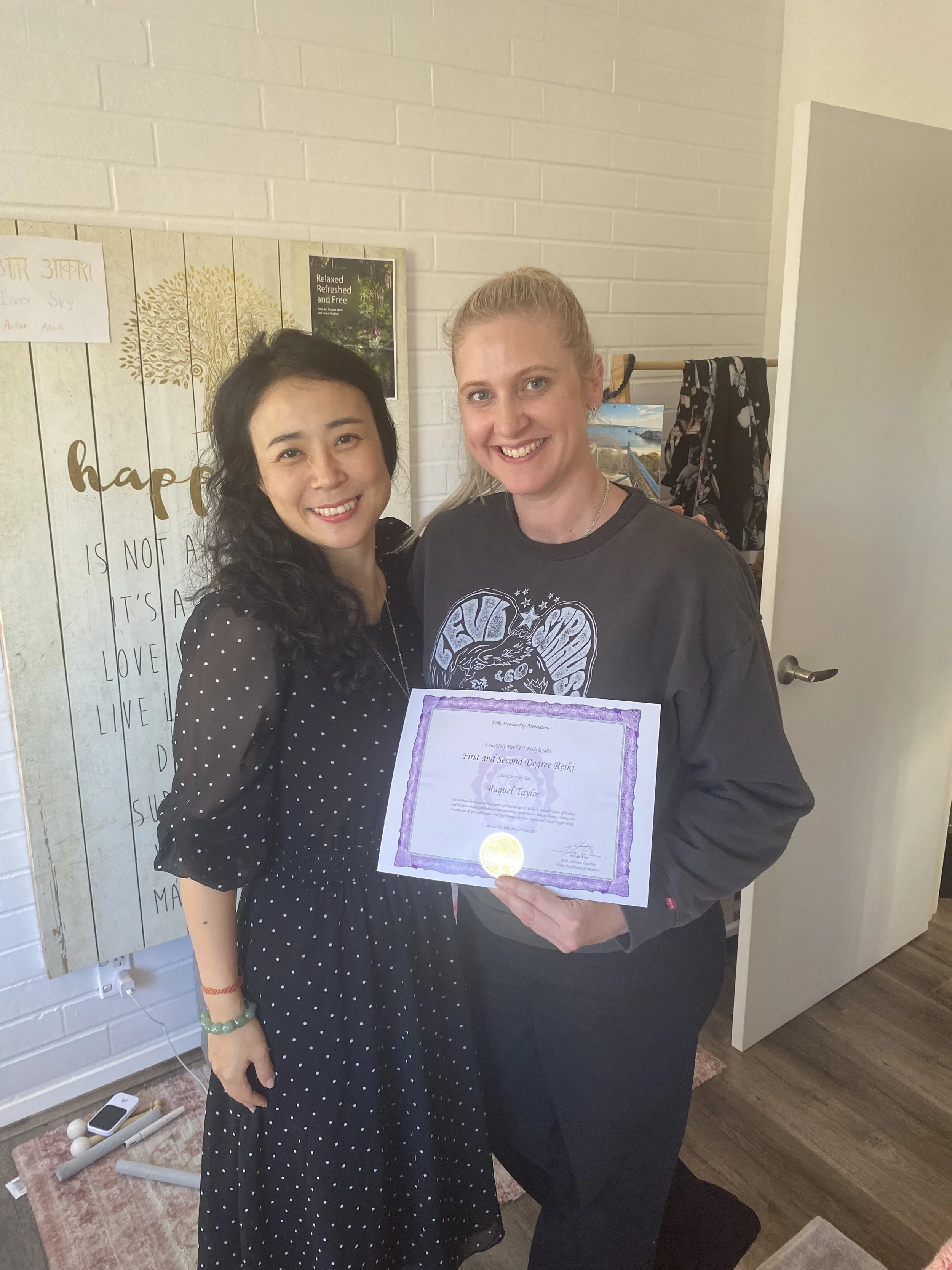 Two women standing indoors, smiling at the camera. The woman on the right is holding a certificate of achievement. The woman on the left has dark hair and is wearing a black dress with white polka dots. The woman on the right has blonde hair tied bac