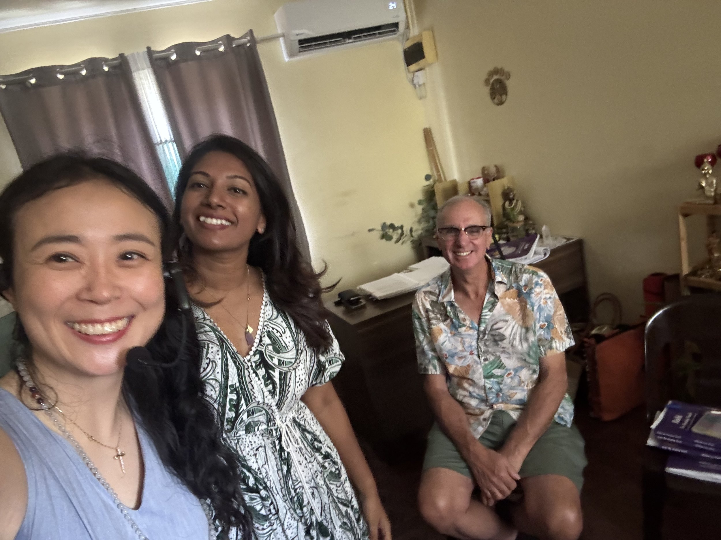 Three people, two women and one man, sitting in a room smiling at the camera. The woman on the left has long dark hair and is wearing a sleeveless gray top. The woman in the middle has long dark hair and is wearing a patterned white and black top. Th