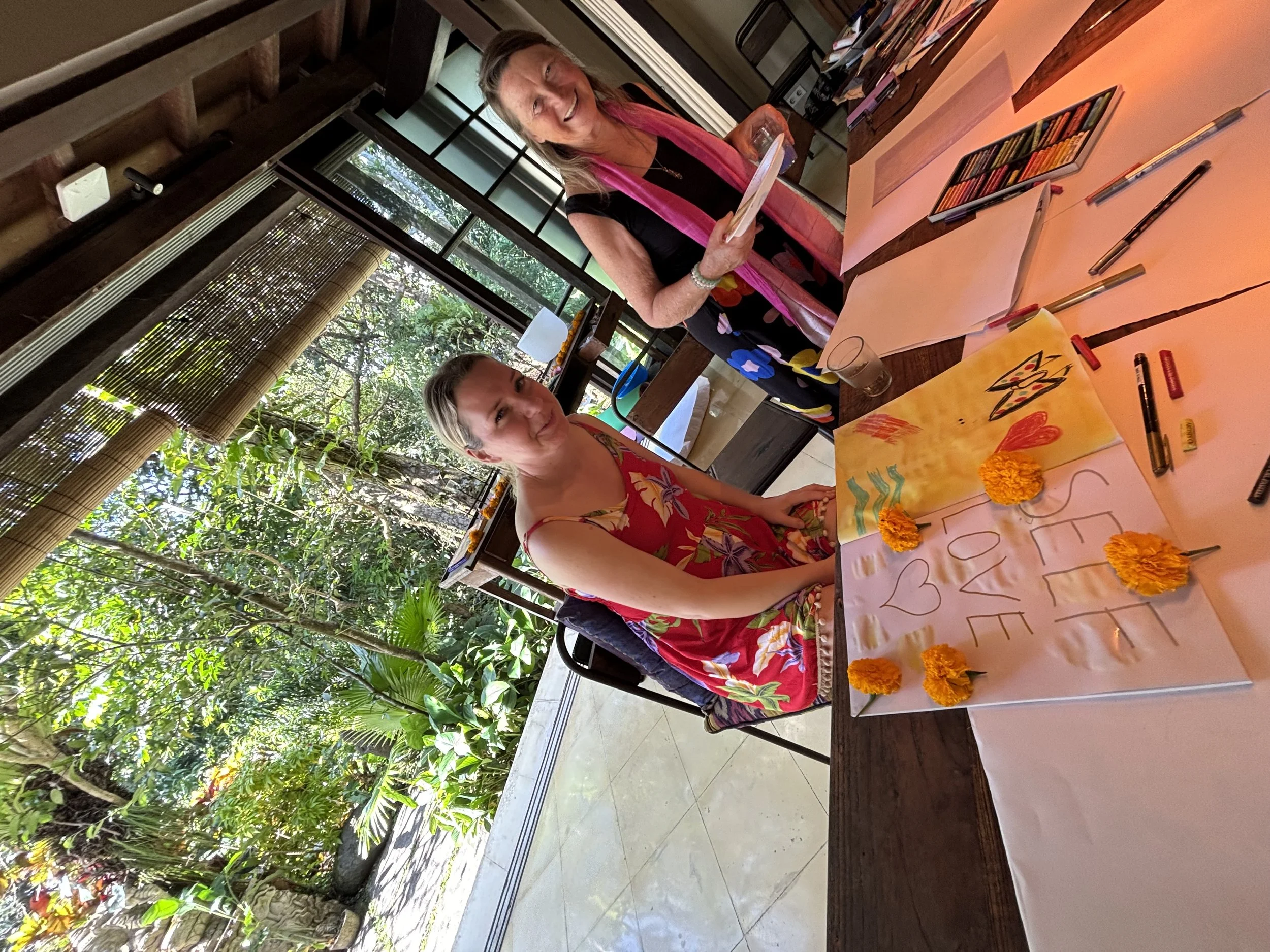 Two women sitting at a table with art supplies and colorful artwork, in a lush outdoor setting.
