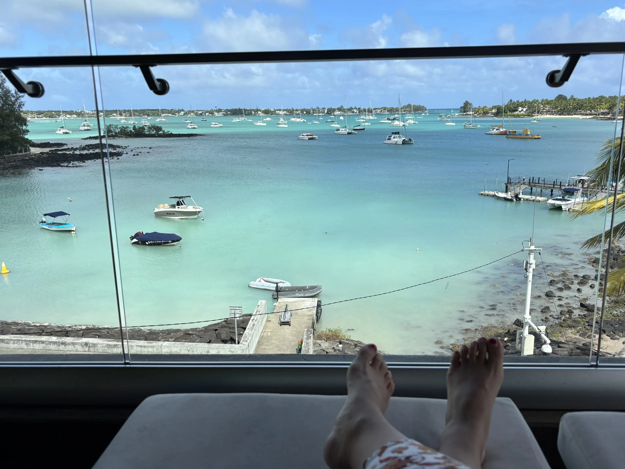 Person relaxing on a lounge chair with feet visible, overlooking a harbor with boats and yachts on turquoise water, a pier, and a clear blue sky with some clouds.