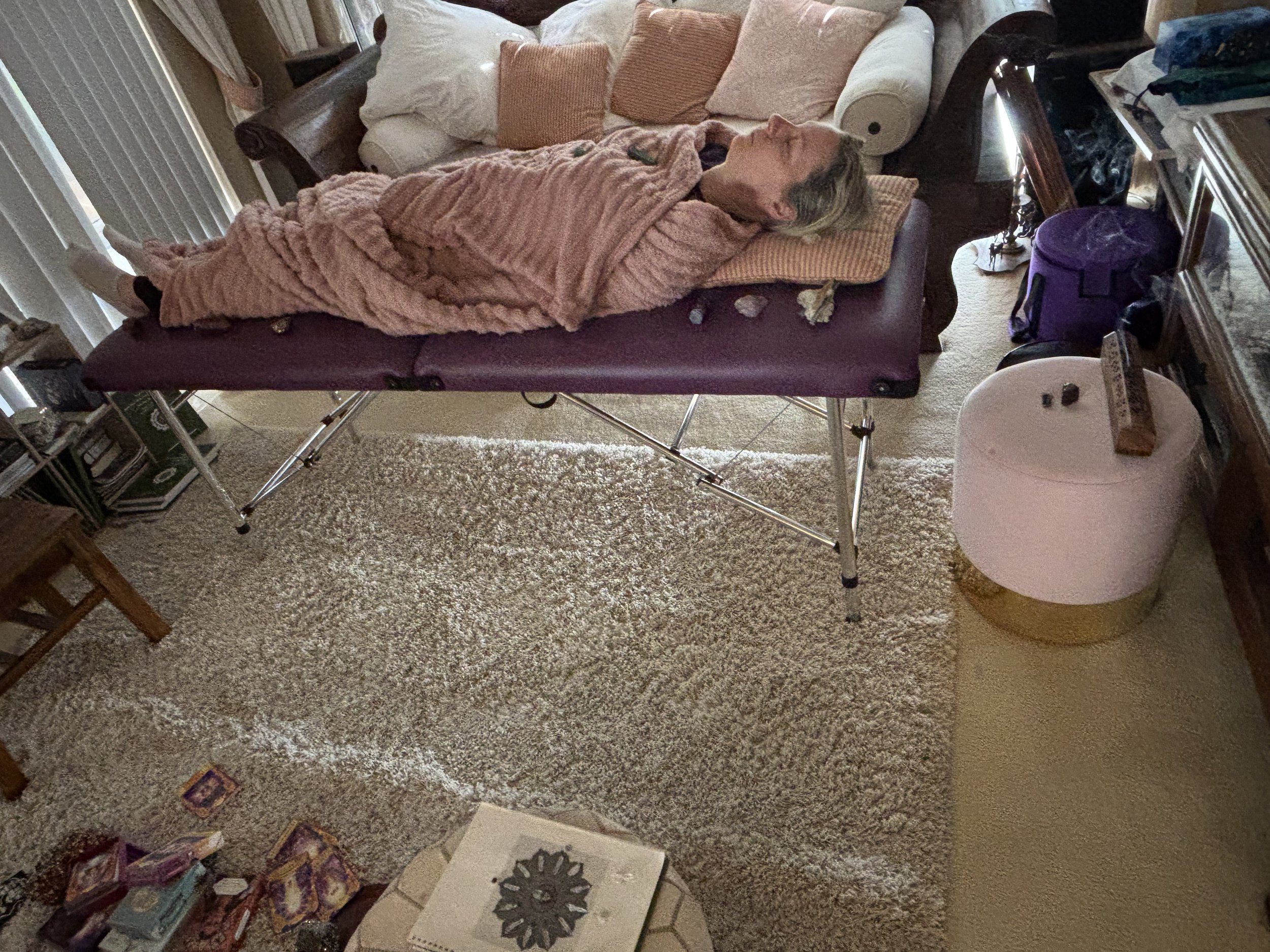 A woman lying on her side on a massage or medical examination table, covered with a pink blanket, in a room with a beige carpet. There are pillows behind her head and on the couch behind her, and various items including books, a purple bag, and a box