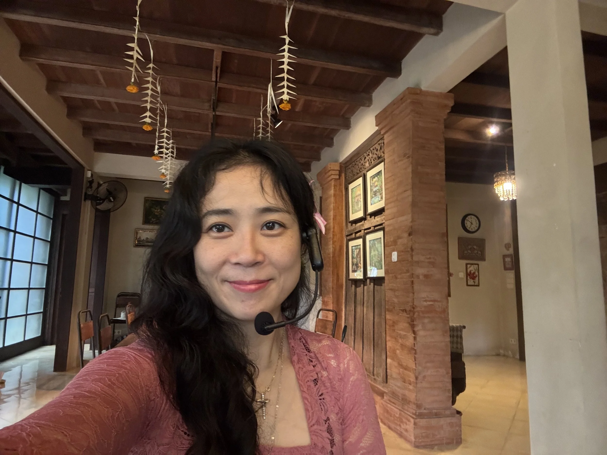 A woman with dark hair wearing a pink top smiling and wearing a headset in a room with wooden beams, hanging decorations, framed pictures, and a large window.