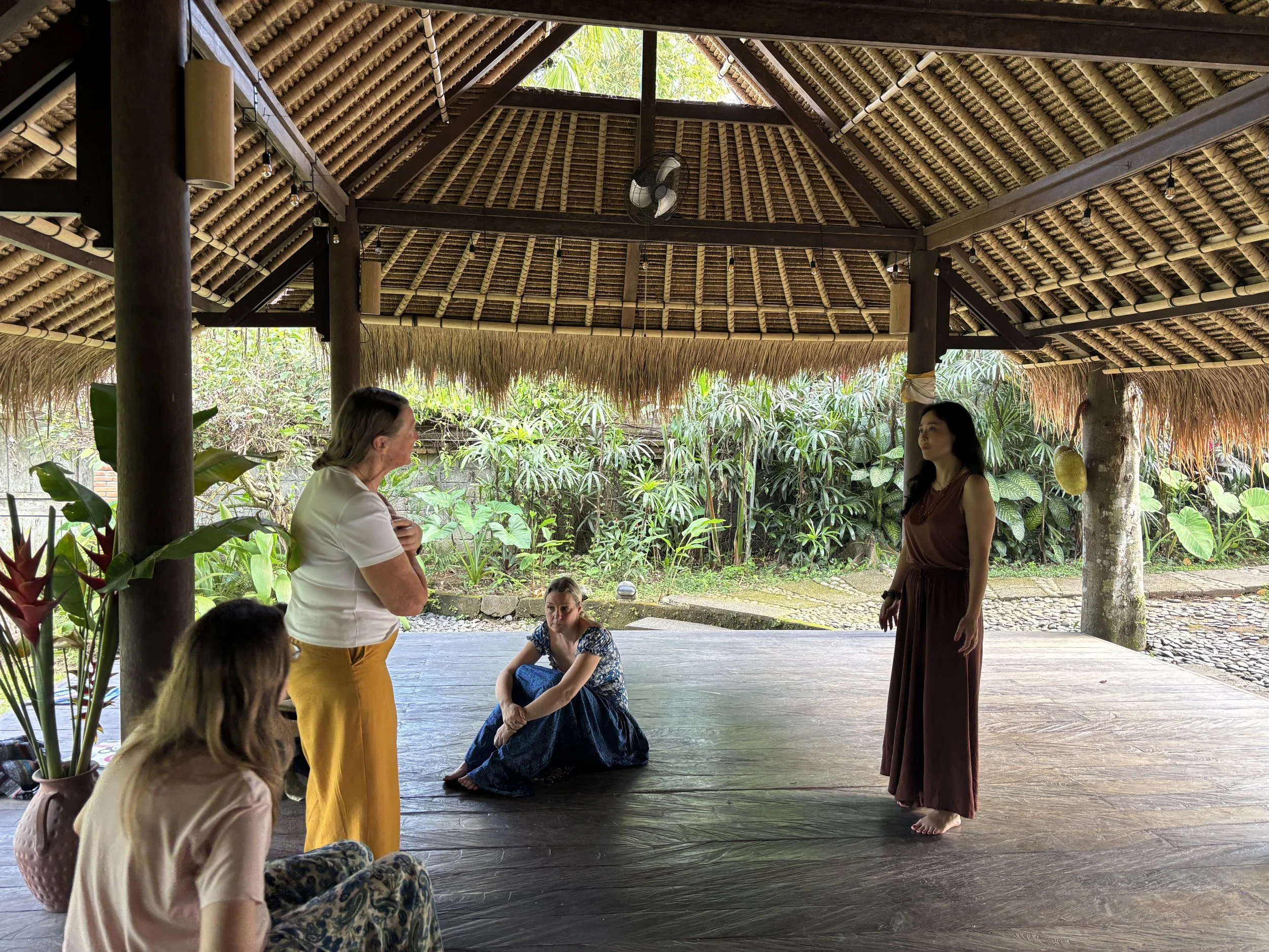 People participating in a group activity or discussion in a thatched-roof outdoor setting with lush greenery.