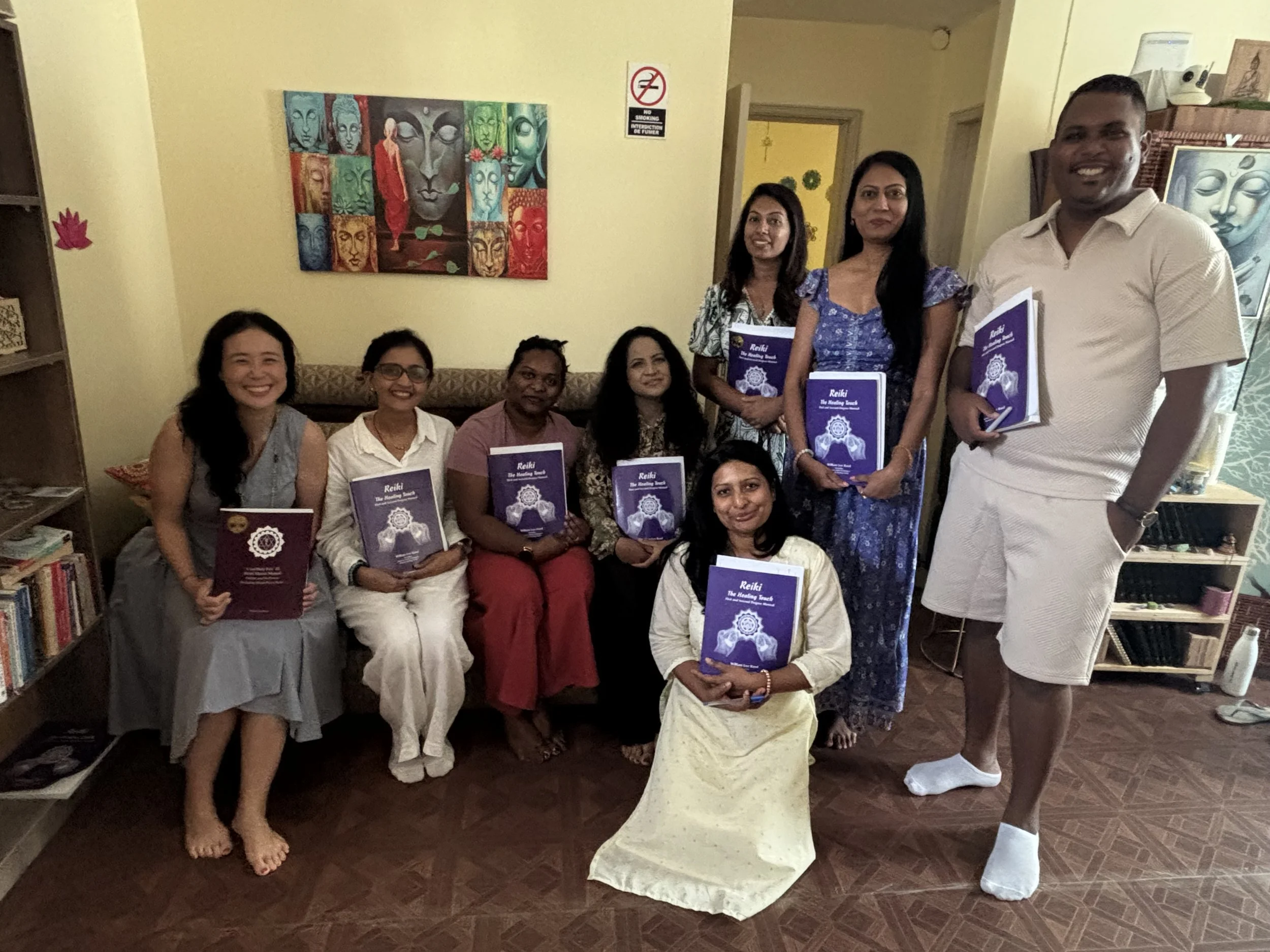Group of nine people, mostly women and one man, gathered indoors holding booklets, smiling for the photo, with artwork and a 'No Smoking' sign on the wall behind them.
