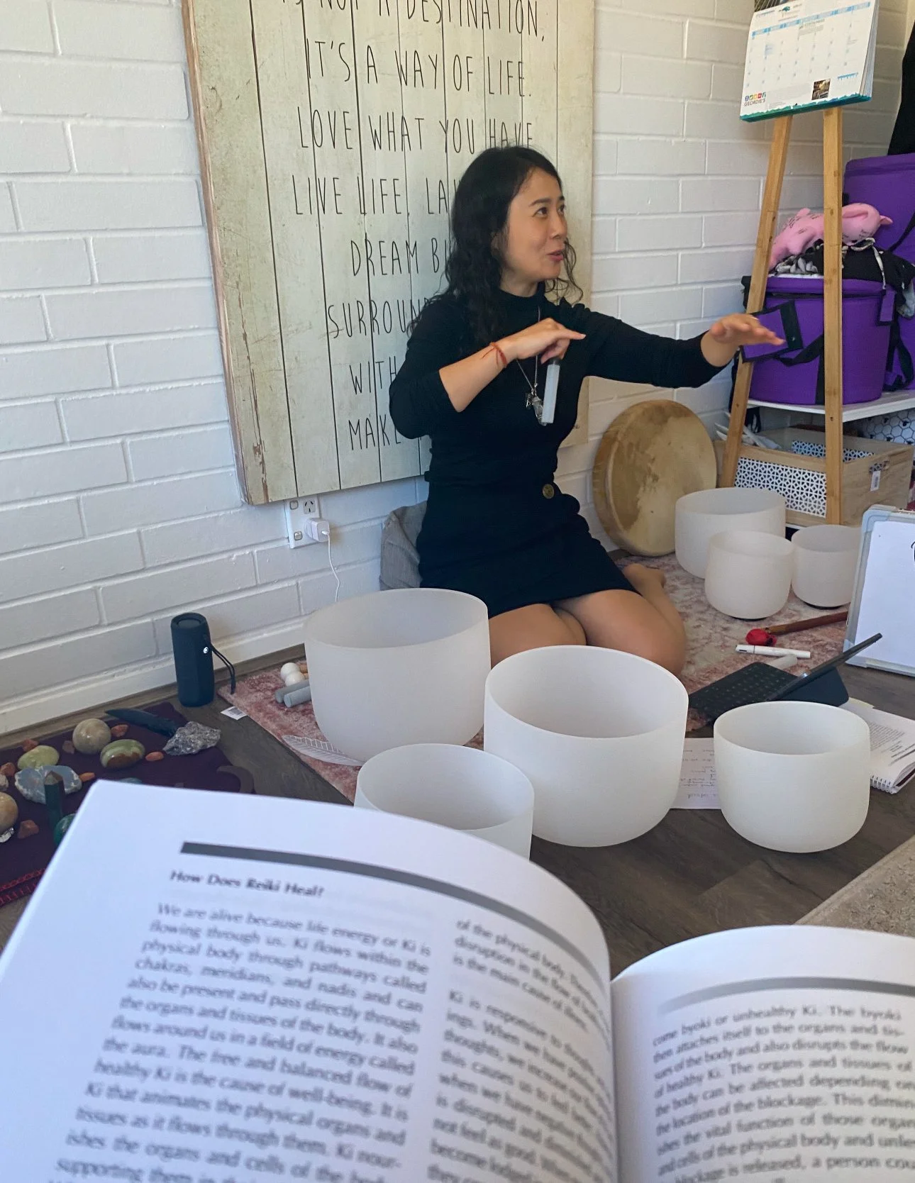 A woman sitting on her knees in front of singing bowls and a music sheet, gesturing with her hands, possibly explaining or demonstrating something, in a room with white brick walls and various objects including a large wooden drum, purple storage bin