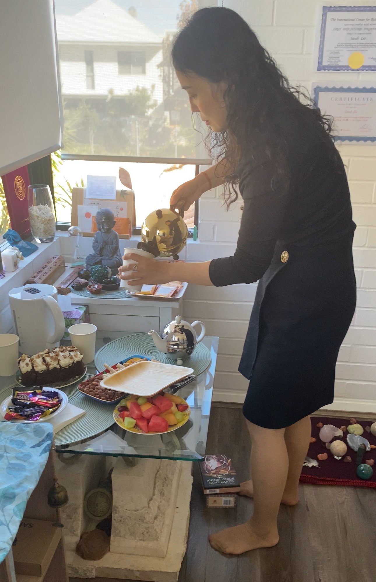 A woman with long dark curly hair, dressed in a black dress with rolled-up sleeves, is pouring a drink from a gold pitcher into a white cup at a table. The table has an assortment of food, including a chocolate cake with whipped topping, a fruit plat