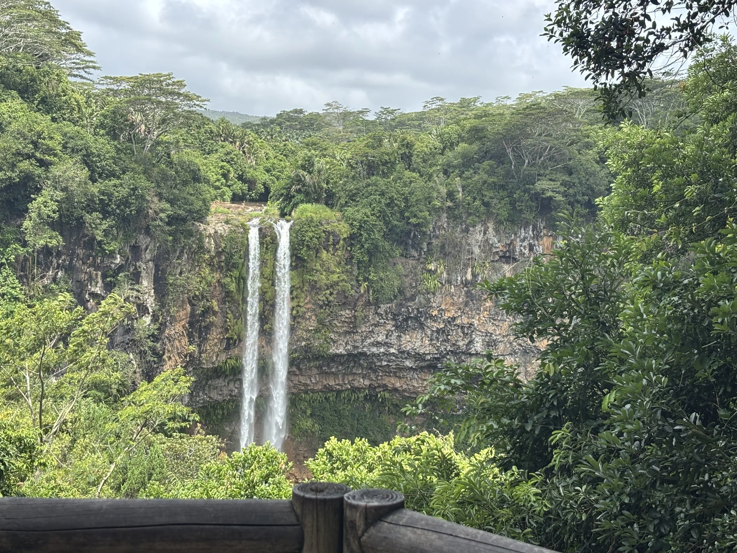 Waterfall cascading down a cliff into a lush green forest, viewed from a wooden railing in the foreground.
