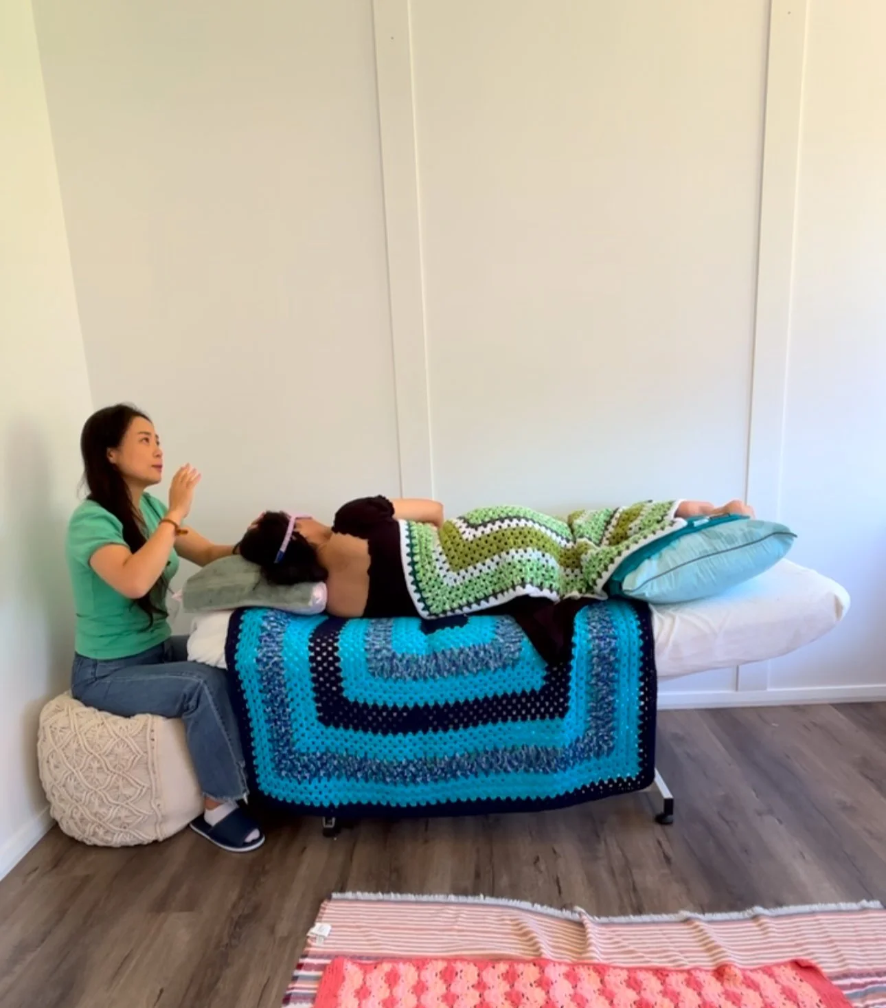 A woman is sitting on a small round cushion next to a table, performing a healing or therapy session with a person lying on a bed covered with a colorful blanket. The person on the bed appears to be receiving treatment, possibly reiki or a similar pr