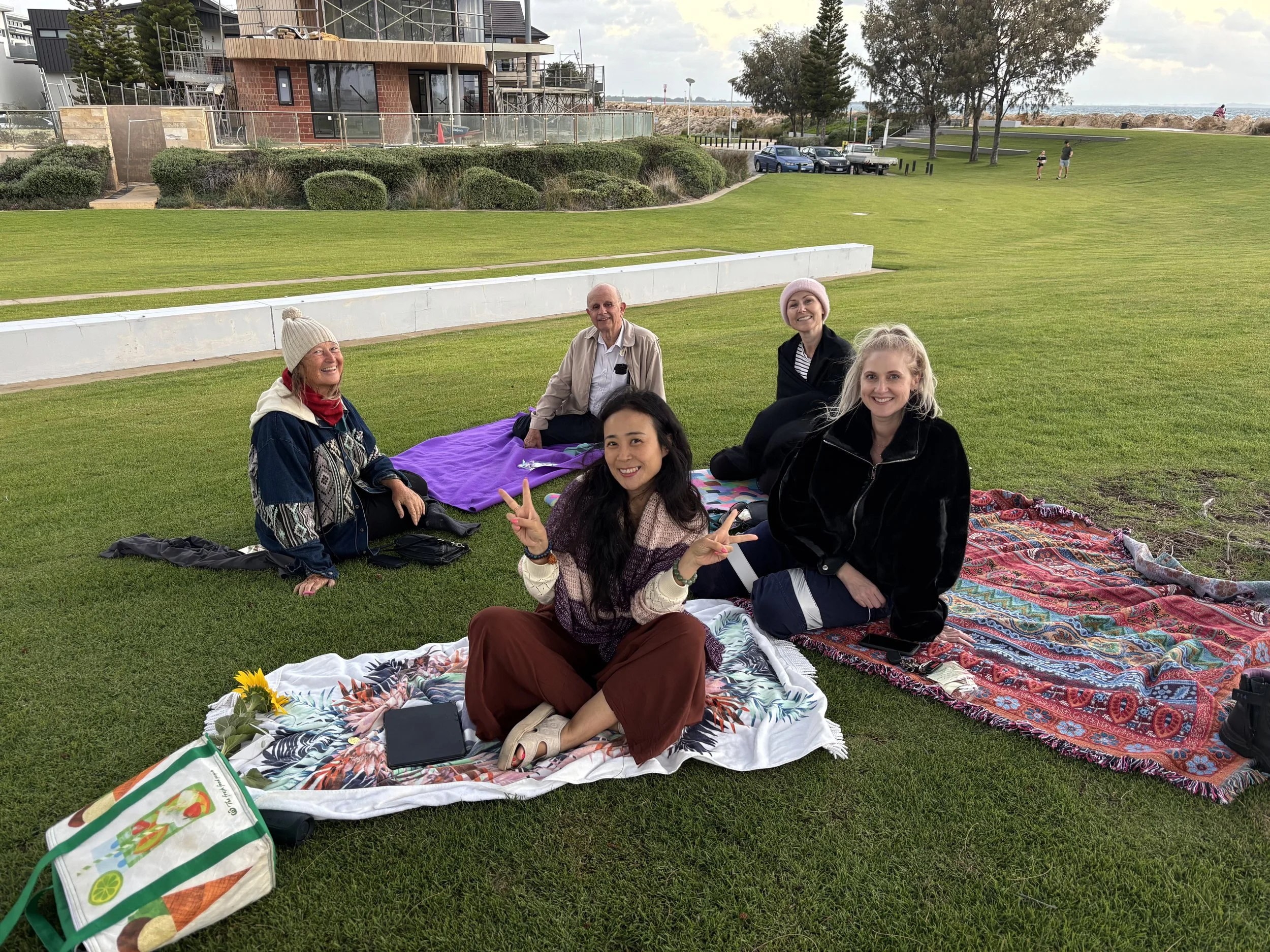 A group of five people sitting on colorful blankets on a grassy area, smiling and posing for the photo, with a building, parking lot, and trees in the background on a cloudy day.