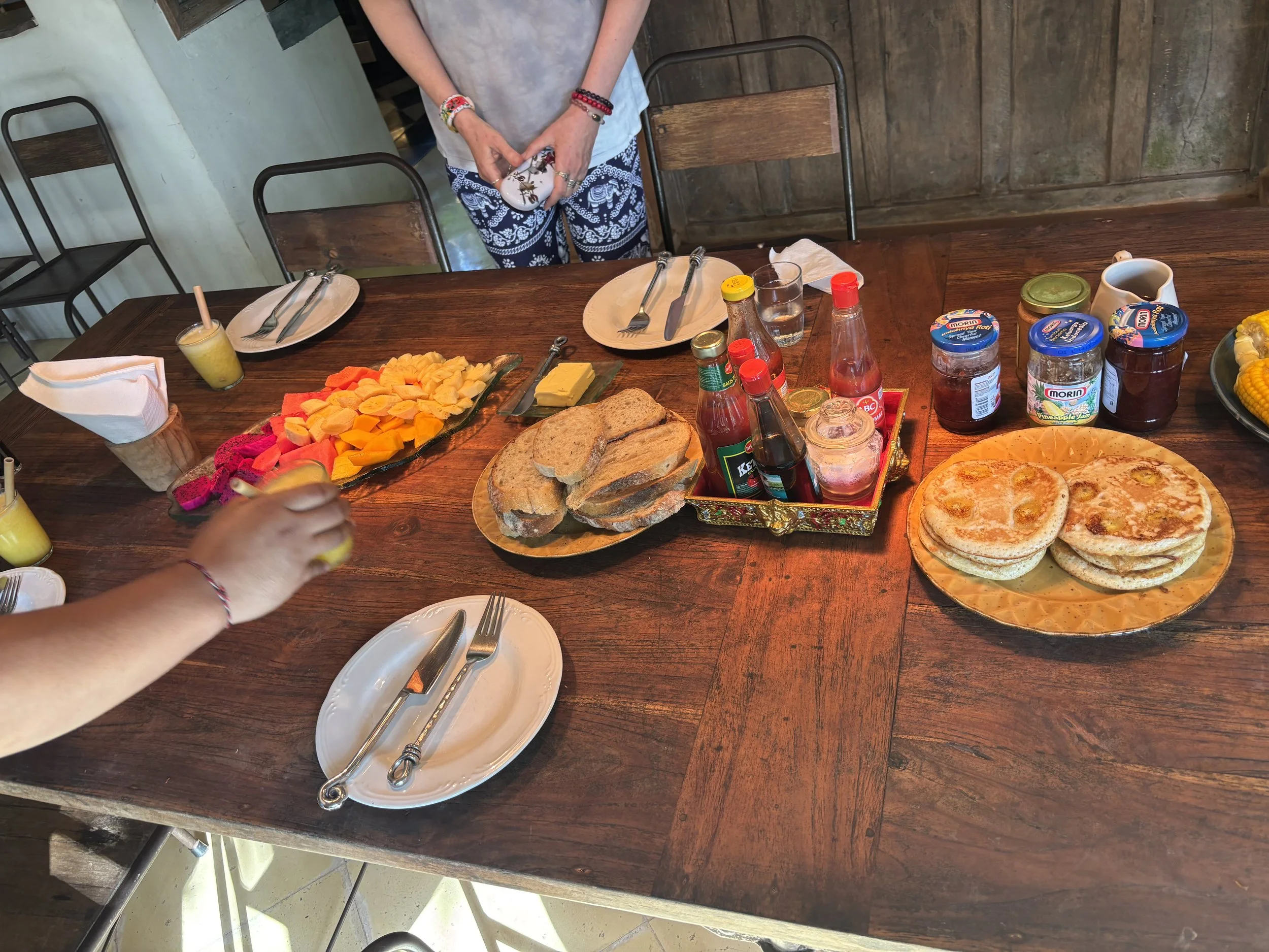 A person standing behind a wooden dining table with breakfast items, including fruit, bread, pizza, and jars of jam, as well as a yellow drink in a glass. The person is holding a small decorative object and has multiple bracelets on their wrists.