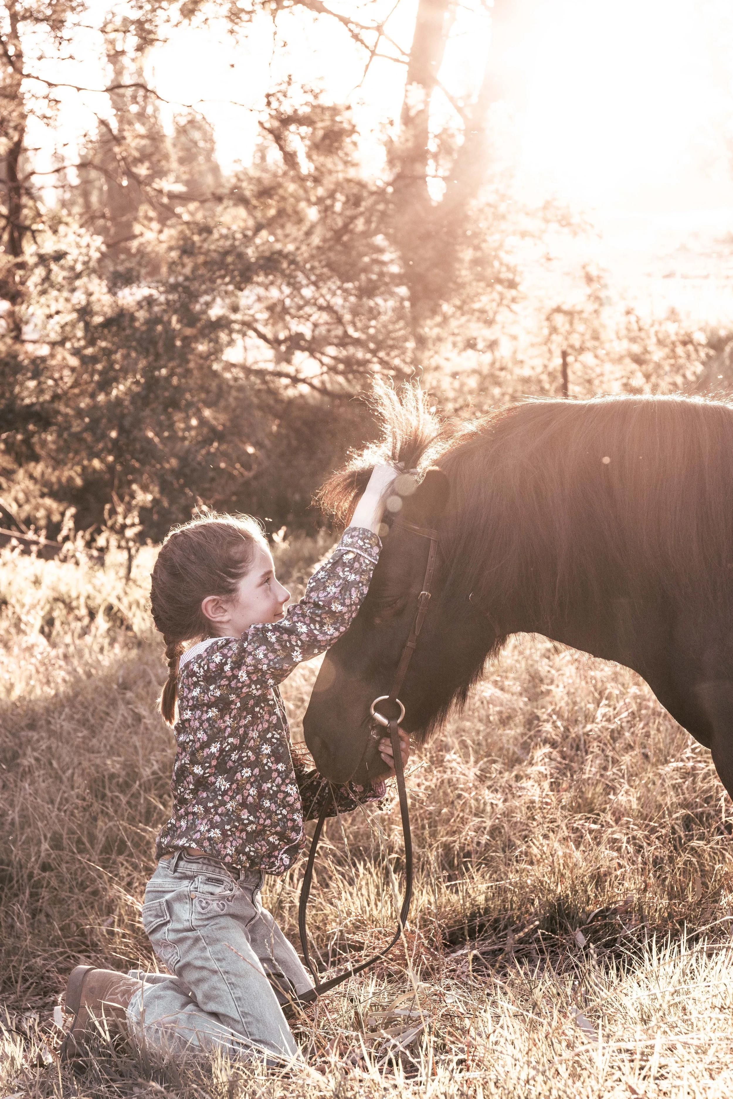 A young girl on the ground playing with a black shetland ponies forelock