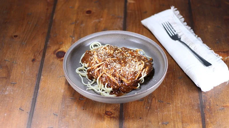 Plate of spaghetti with meat sauce and grated cheese, with a fork on a white cloth napkin beside it on a wooden table.