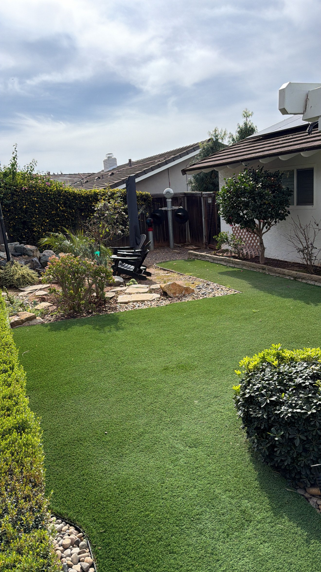 A backyard garden with a lush green lawn, shrubbery, and a small rock garden with stepping stones. There are two black Adirondack chairs, a patio umbrella, and a weather station near the back fence. The background includes a house with a chimney and a partly cloudy sky.