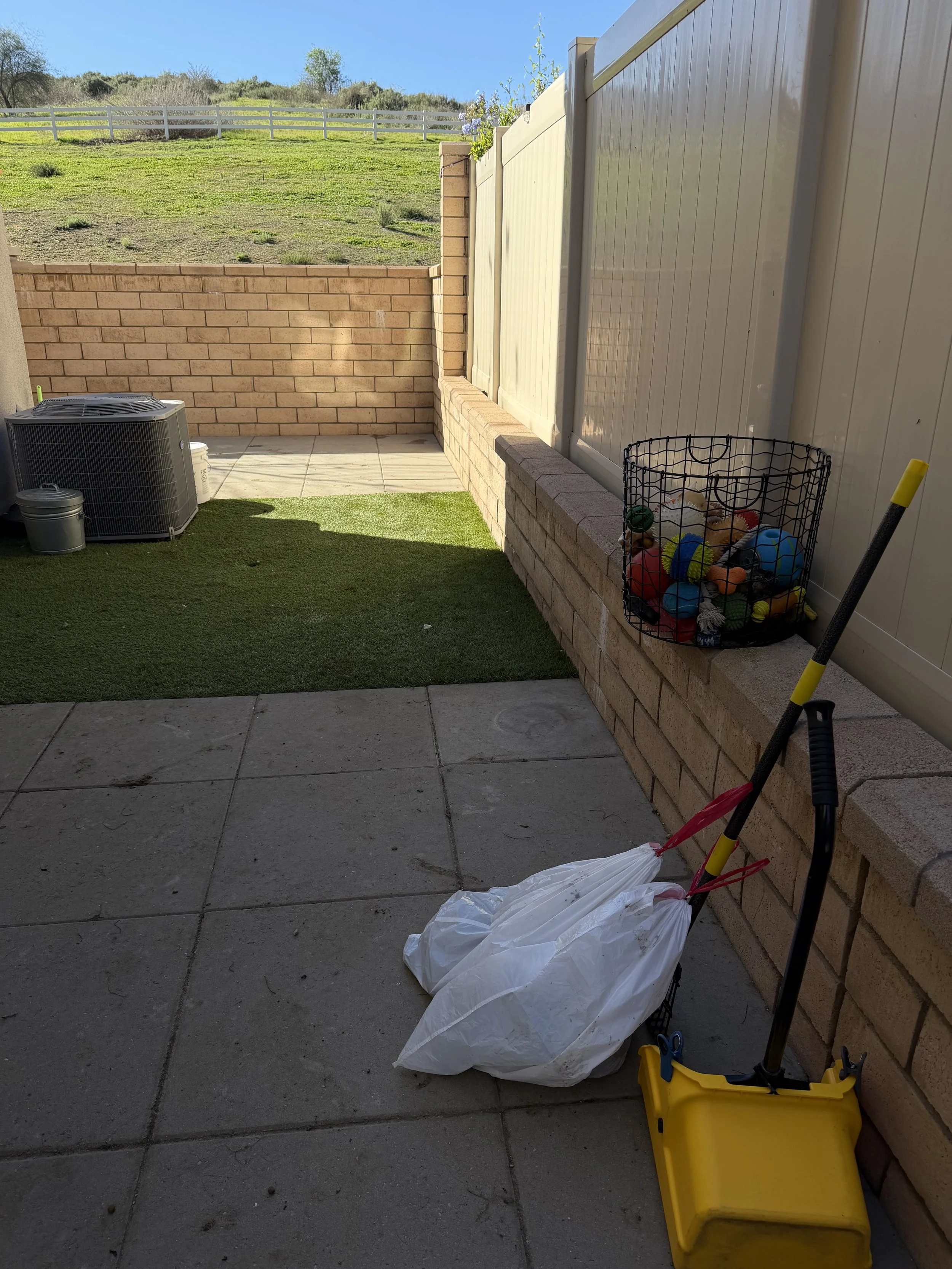 Backyard patio with artificial grass, a basket of sports balls, a yellow broom, and an air conditioning unit on a concrete pad, with a brick wall and a fenced grassy hill in the background.
