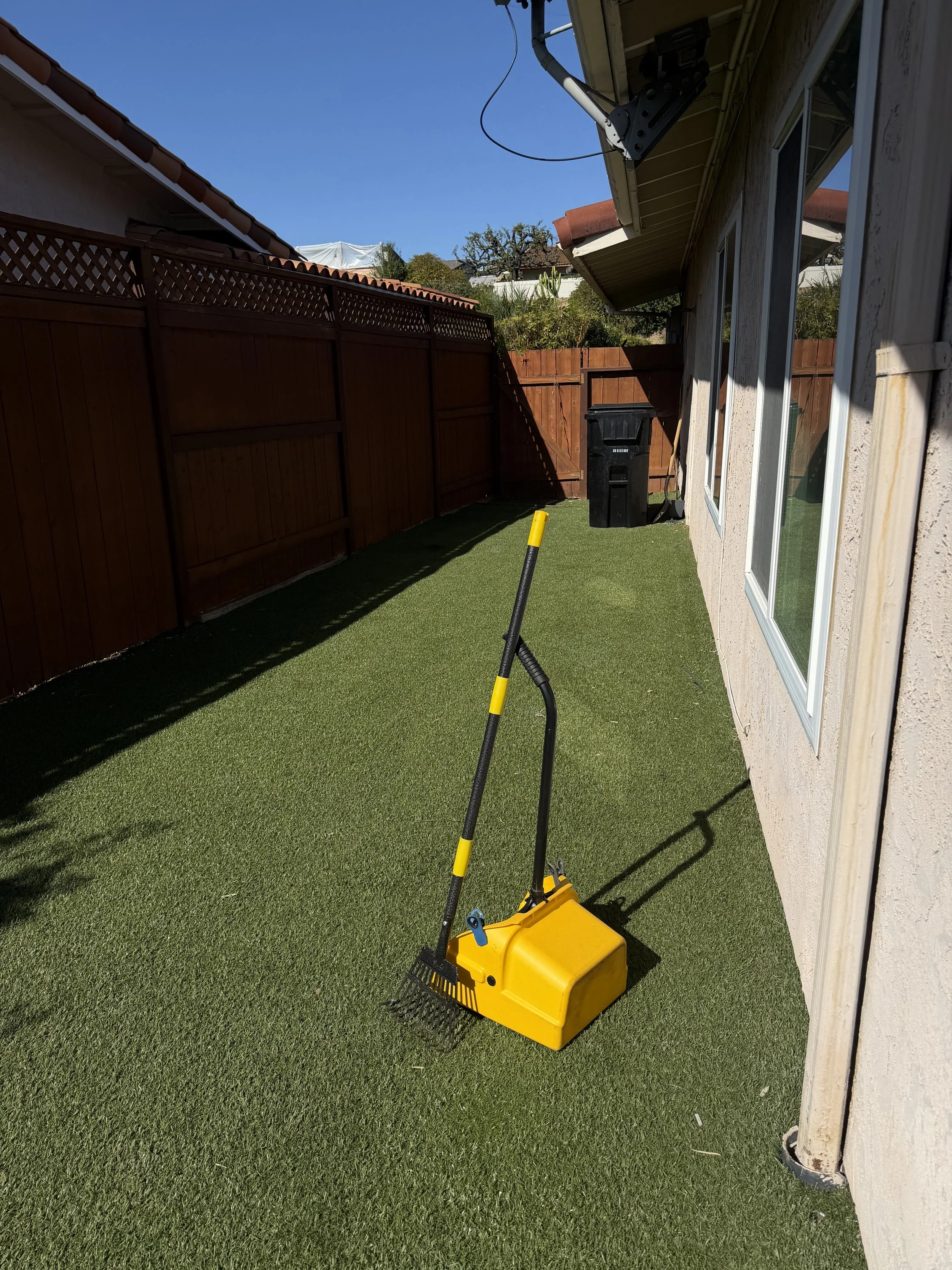 A yellow backyard leaf blower with a black handle on synthetic grass next to a house wall with windows, a black trash bin, and a wooden fence in the background.