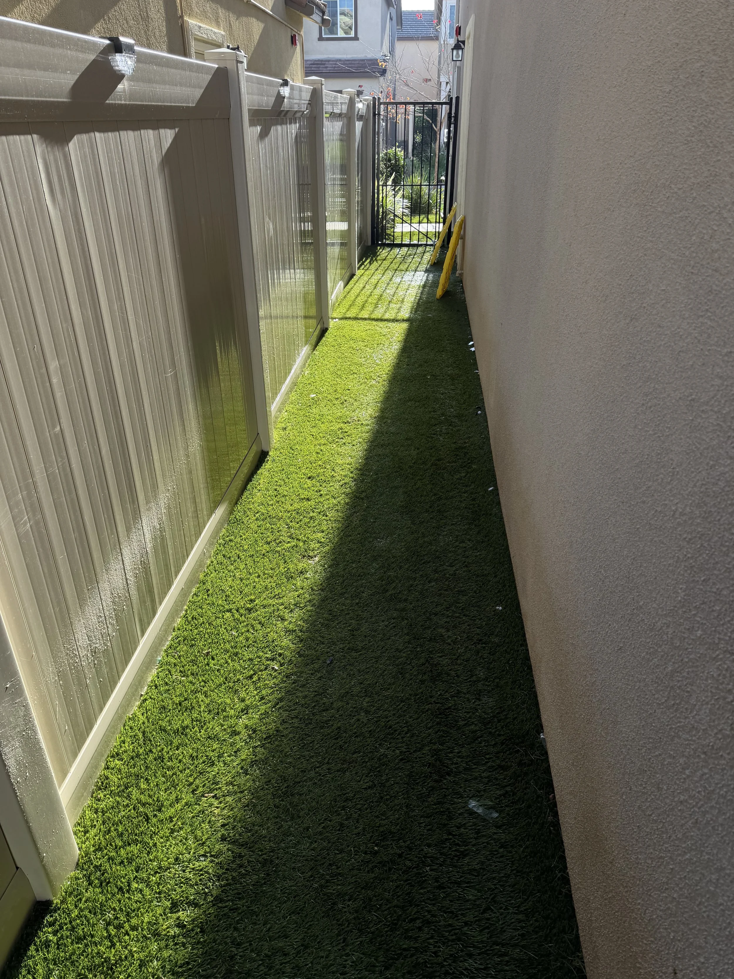 Narrow outdoor pathway with artificial green grass, enclosed by a beige wall on the right and a beige fence on the left, leading to a black metal gate with plants visible beyond.