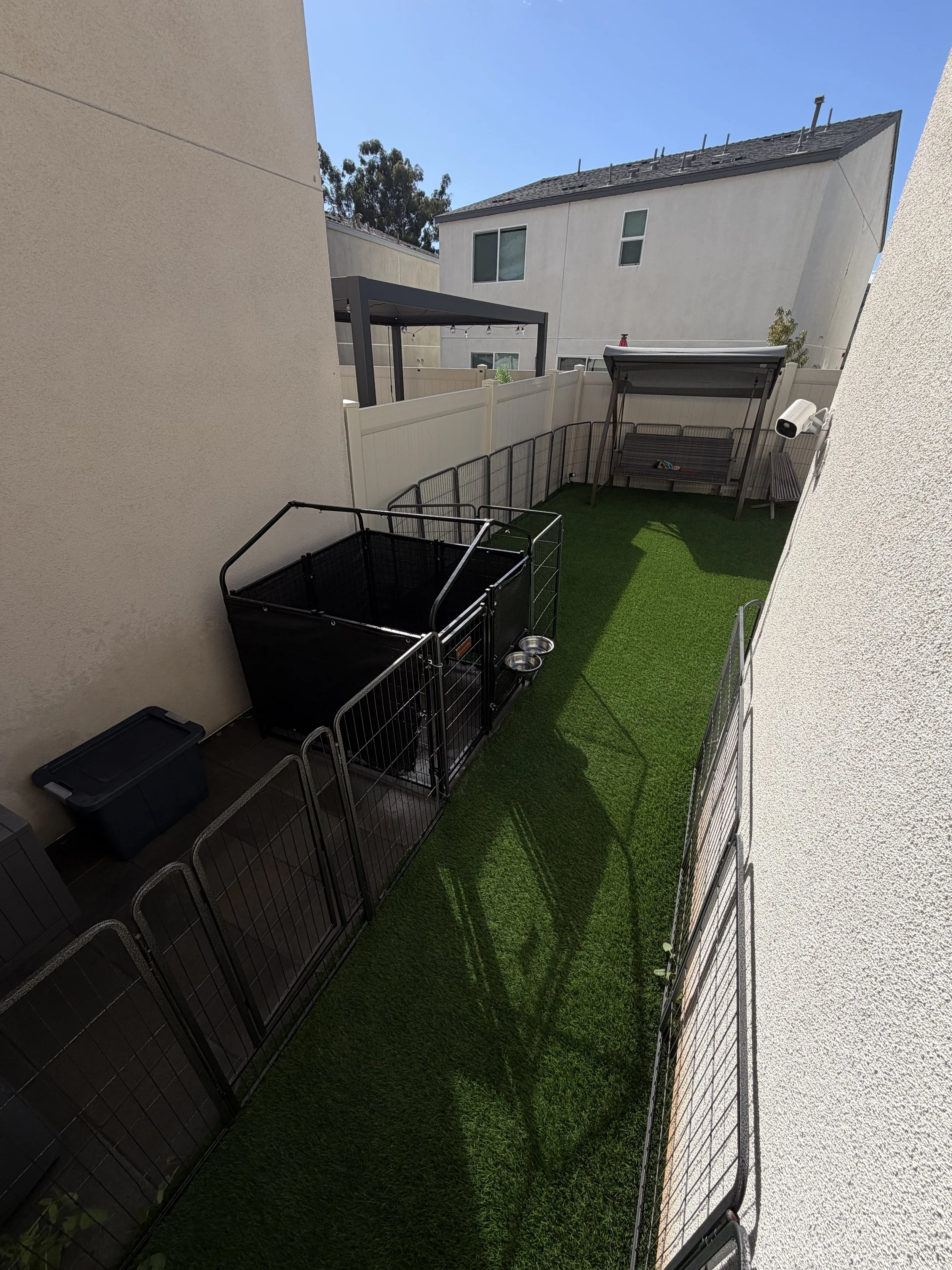 Small backyard with artificial grass, enclosed by a white vinyl fence. Contains a black pet crate, a metal dog pen, a gray swing with a canopy, and a bench. Shadows of plants are cast on the grass.