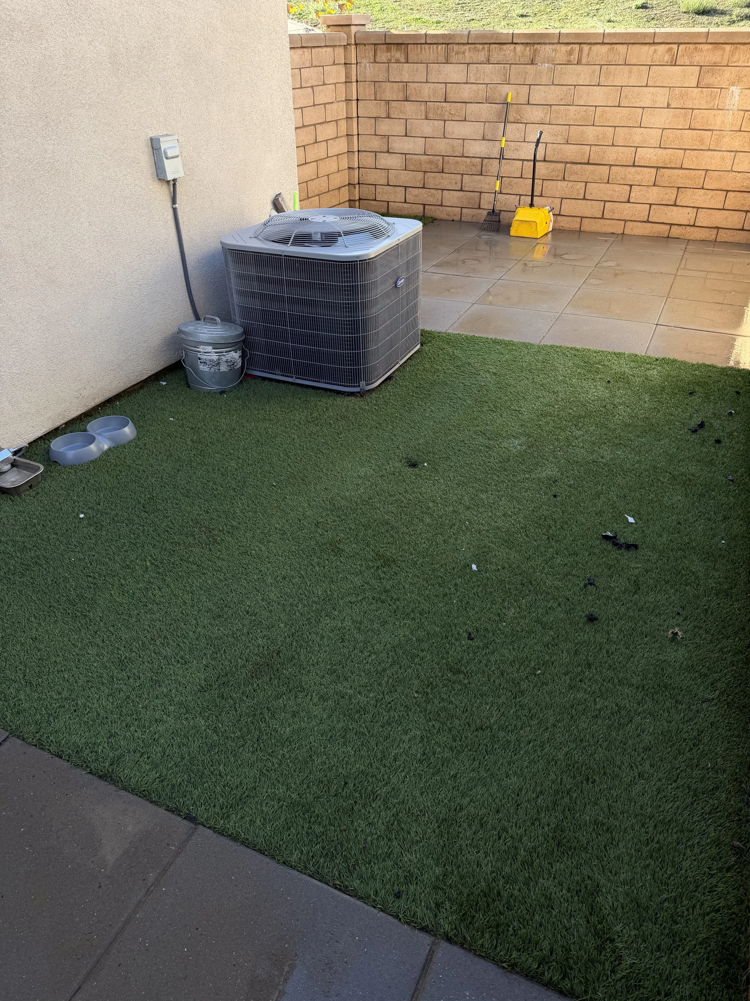 Backyard with artificial grass, a large air conditioning unit, a bucket, two pet bowls, and cleaning tools on a tiled patio area.