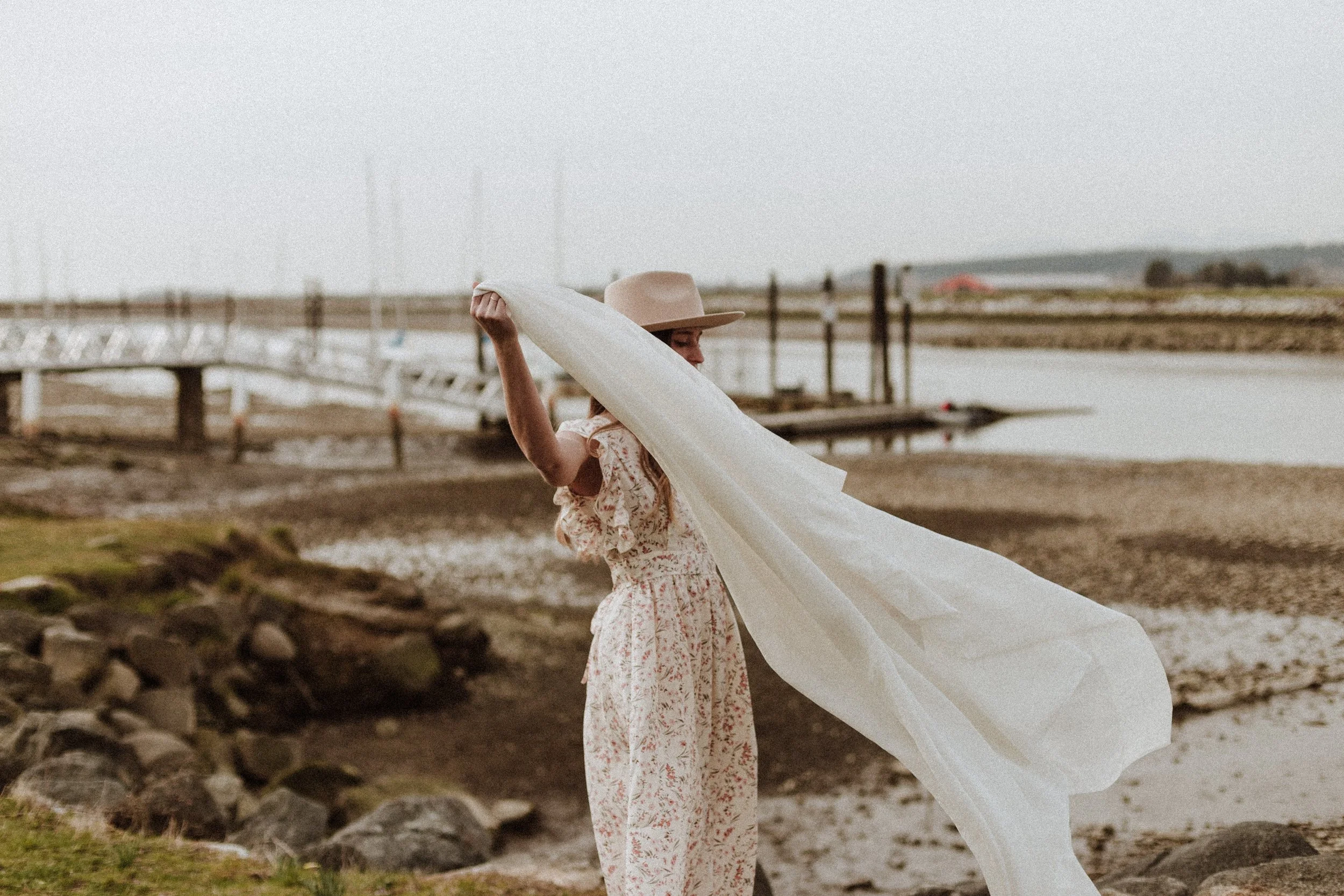 A woman wearing a floral dress and a wide-brimmed hat stands on a rocky lakeside, holding a long flowing white fabric that billows in the wind, with docks and boats in the background under an overcast sky.