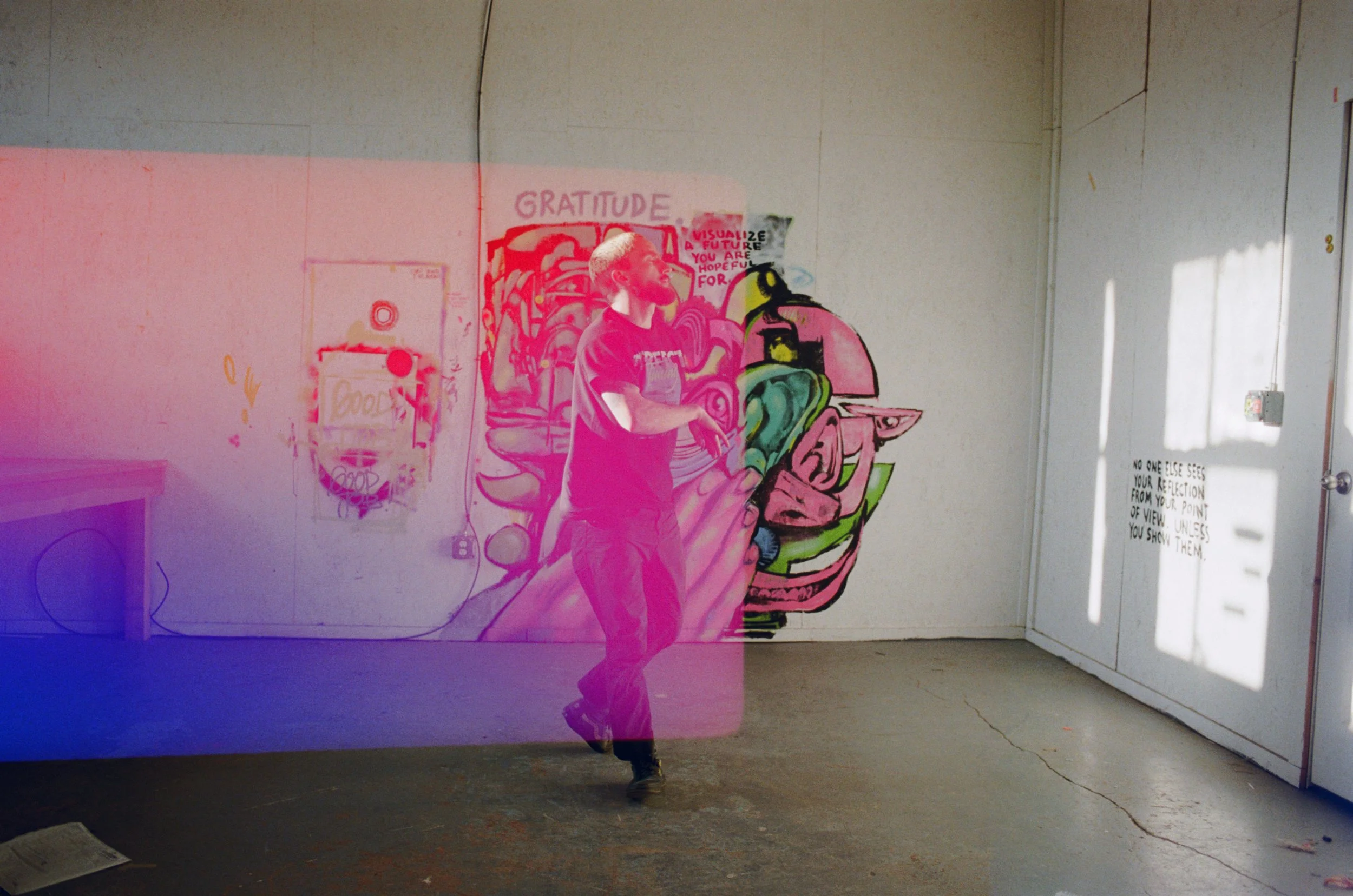 A person with a beard wearing a black T-shirt and jeans in front of a colorful  mural on a white wall. The mural features abstract and cartoonish elements, including a large face and other designs, with the word 'GRATITUDE' written at the top. The room has a concrete floor and a door to the right with sunlight casting shadows. There is also a small sign with text to the right side of the door.