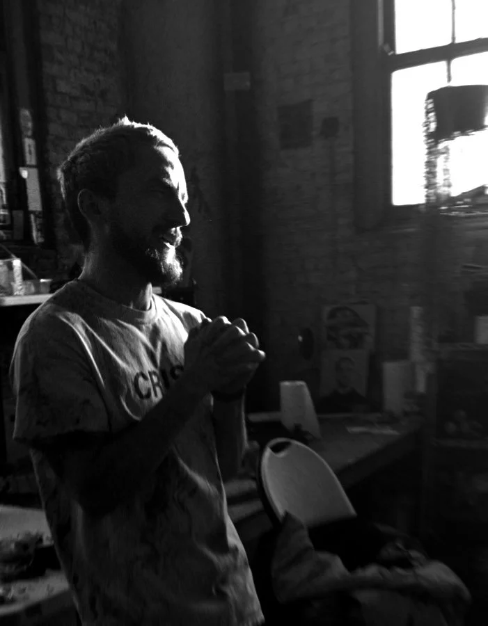 Man with a beard and short hair, smiling and clasping his hands, standing in a dimly lit room with brick walls and a window in the background.