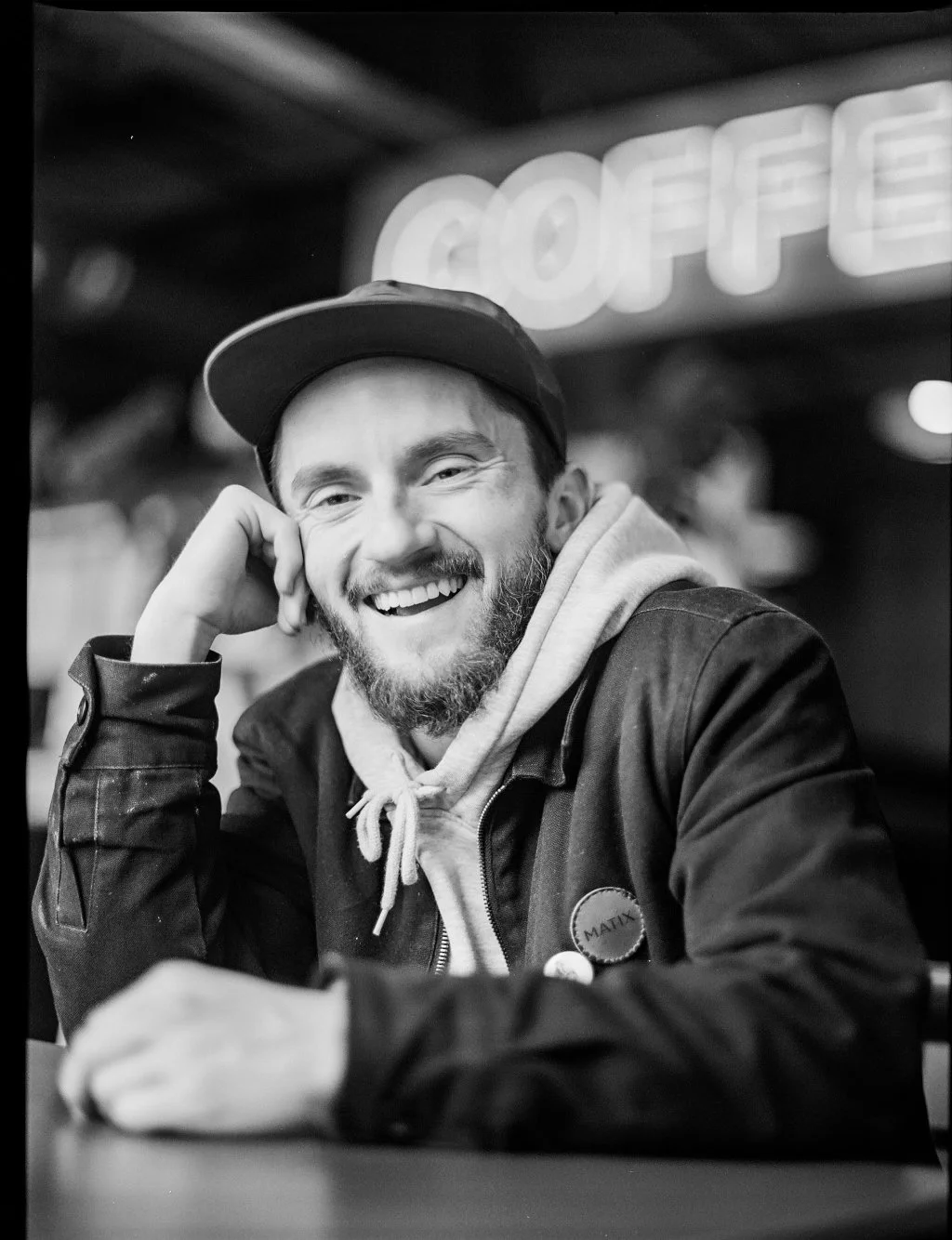 Black and white photo of a smiling man with a beard, wearing a baseball cap, hoodie, and jacket, sitting at a table in a casual setting with a neon sign in the background.
