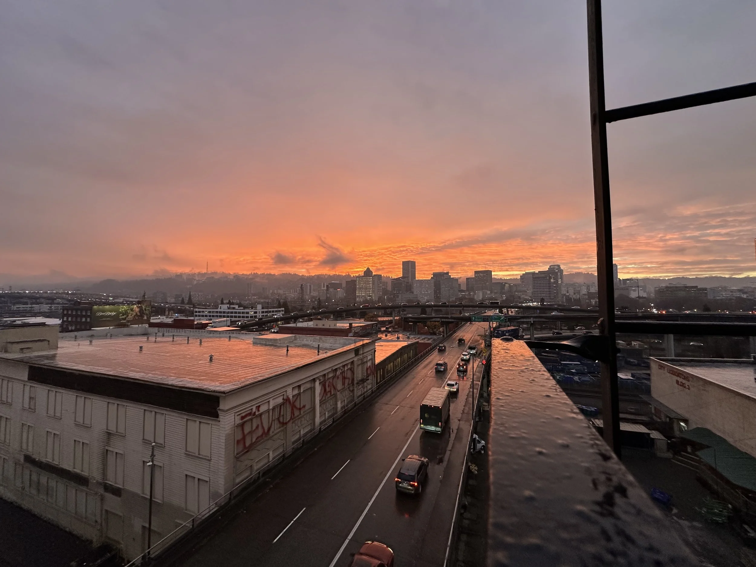 Cityscape at sunset with orange and pink clouds, buildings in the distance, a highway with cars, and a rooftop with graffiti.