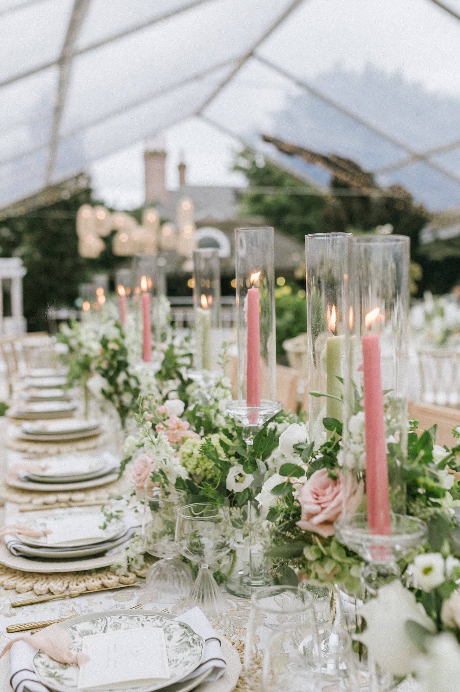 Elegant outdoor banquet table setting under a tent, with tall candleholders, pink and white flowers, plates, and glasses.