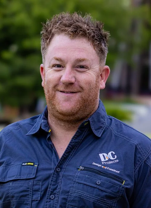 Smiling man in a blue shirt with "DC Projects" logo outdoors.