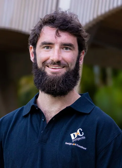 Man with a beard wearing a black polo shirt with a 'Design and Construct' logo, standing outdoors with greenery in the background.