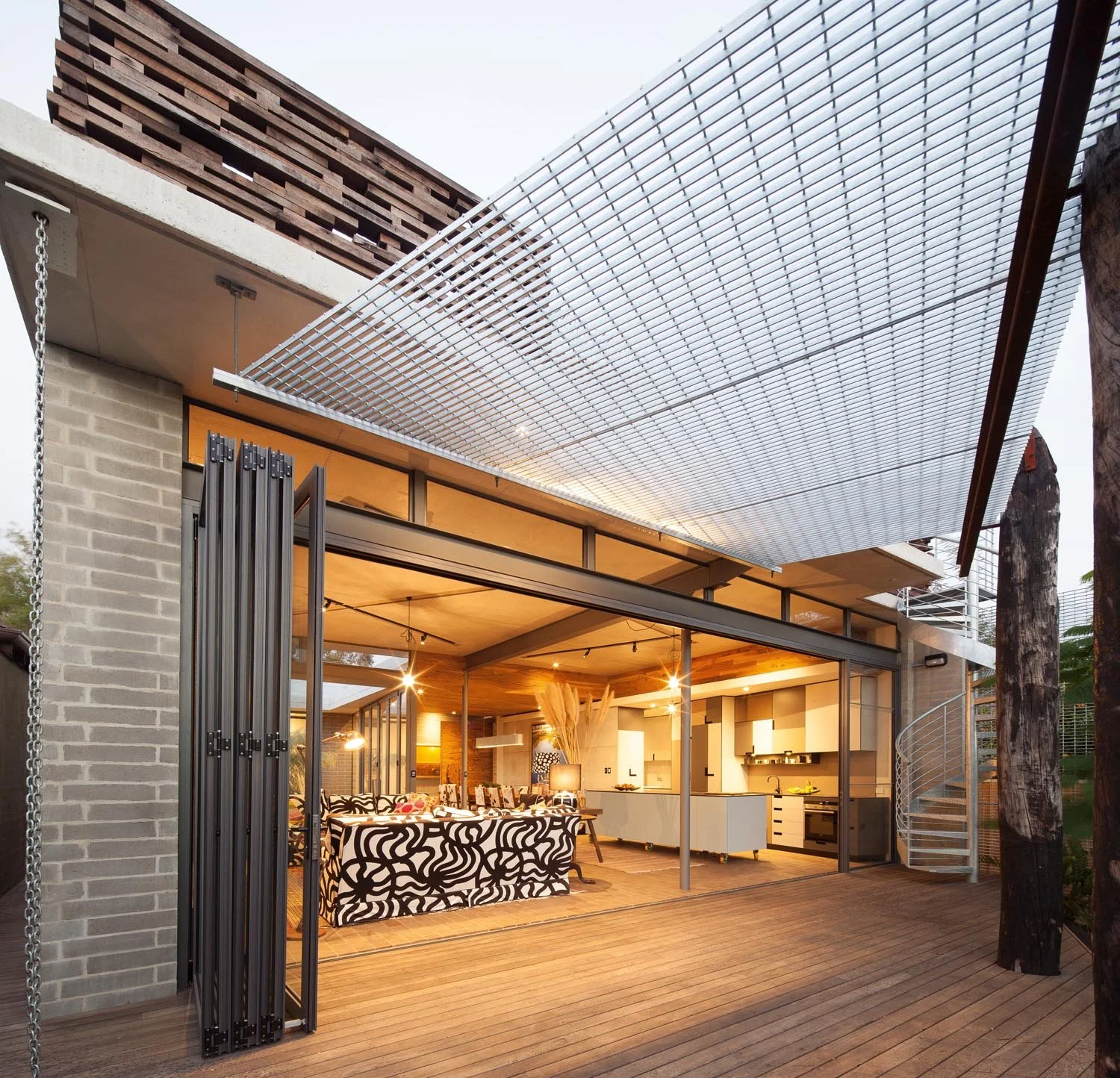 Modern open-plan kitchen and dining area with sliding glass doors, featuring contemporary furniture, a wooden deck, spiral staircase, and a grid-like metal canopy.