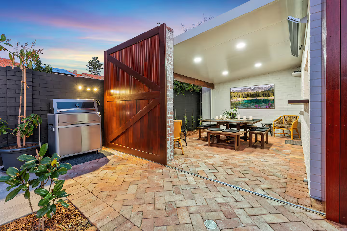 Outdoor patio with a modern grill, wooden dining table, chairs, potted plants, brick flooring, and a large wooden gate.