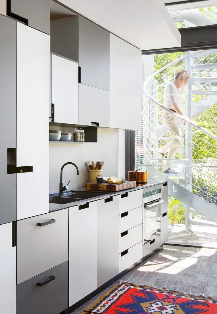 Modern kitchen interior with sleek white and black cabinets, a black countertop with a sink, a colorful geometric rug, and a spiral staircase visible through large windows.