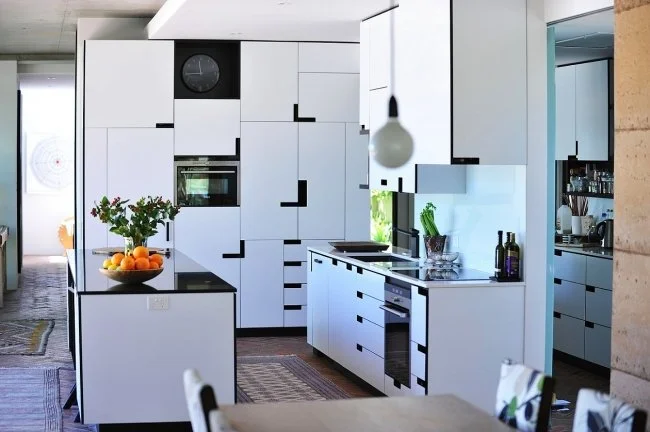 Modern kitchen with minimalist design featuring white cabinets with black accents, stainless steel appliances, and a black countertop. A vase with flowers and a bowl of oranges sit on the island. Bright and clean space with a subtle contemporary aest