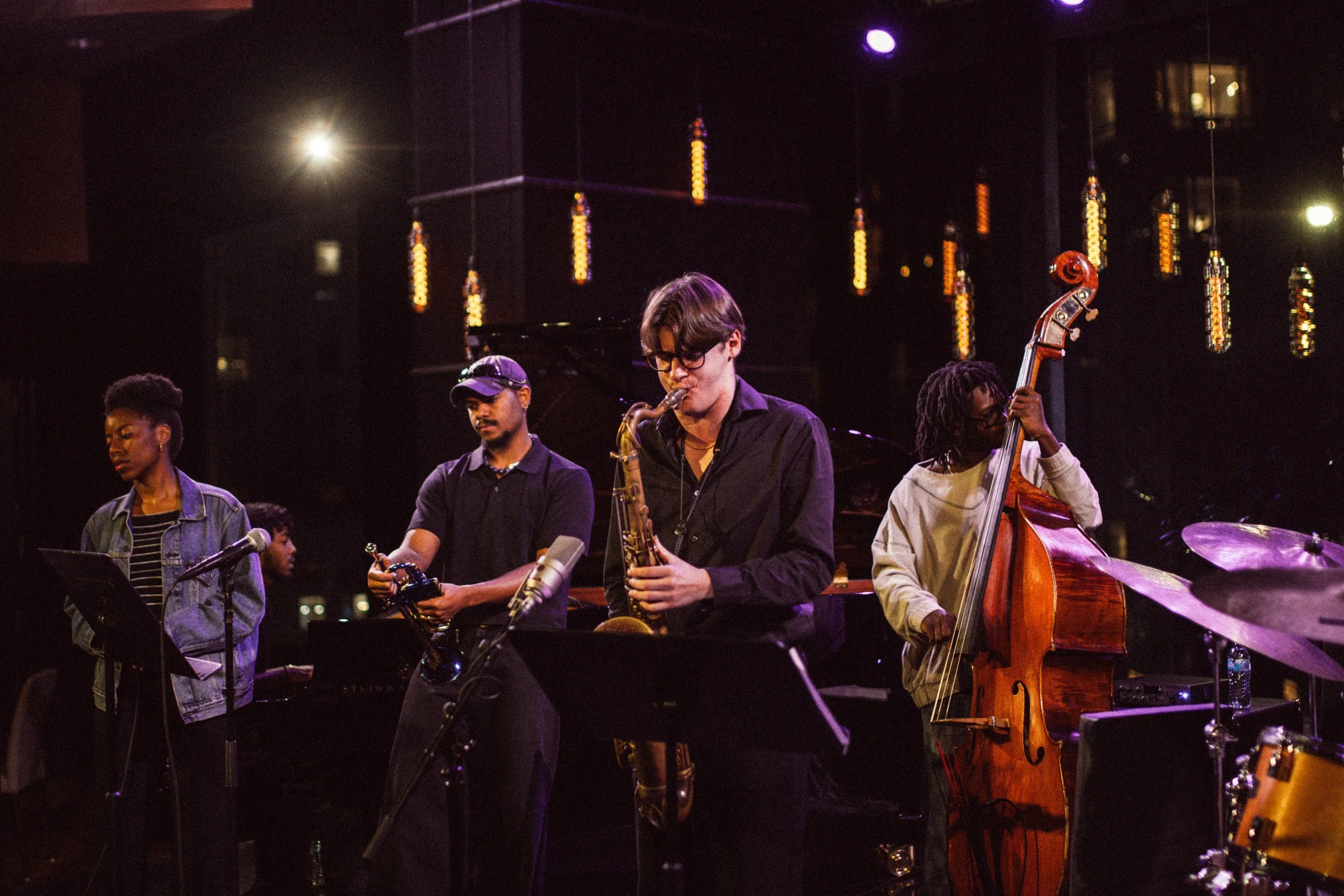 A group of four musicians performing on stage at a jazz concert. The musicians include a female vocalist, a saxophonist, a double bassist, and a drummer. The stage is dimly lit with colorful lights and a dark background.