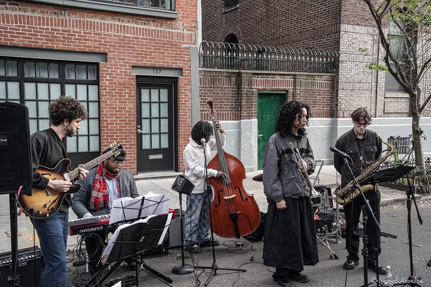 A street band performing outdoors with five musicians playing various instruments including a guitar, keyboard, double bass, and saxophone, in front of brick buildings.