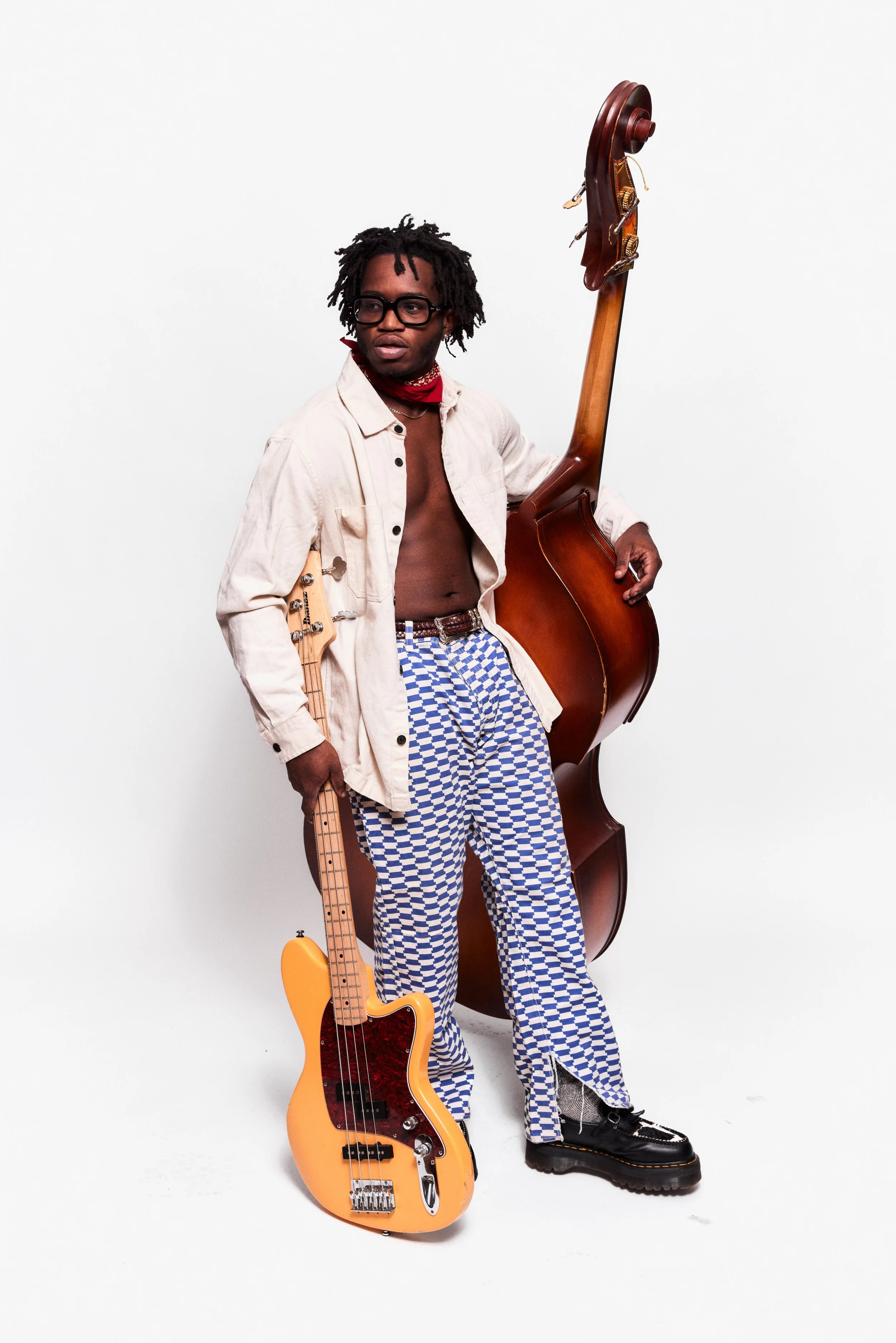 A man with dreadlocks and glasses holding a yellow electric bass guitar. He is dressed in a light-colored shirt, blue and white checkered pants, and black shoes. He stands next to a large upright double bass, against a plain white background.